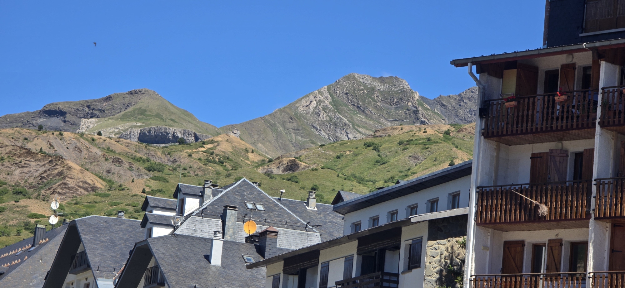 The image shows a view of the town of Sallent de Gállego, Spain, with a backdrop of rugged mountains under a clear blue sky. In the foreground, modern buildings with balconies and dark pitched roofs are visible. One building has a white facade with multiple floors, adorned with wooden shutters and railings on the balconies. A broom is propped against a railing on one of the balconies. The mountainous landscape behind the town features steep, green slopes interspersed with rocky outcrops and patches of dry grass. The lower slopes of the mountains show signs of erosion and varied vegetation. The sky is a vibrant, cloudless blue. A small bird is visible in flight in the upper left portion of the image. The scene depicts a typical day in a mountain town, likely during a warm season given the green vegetation and clear sky.