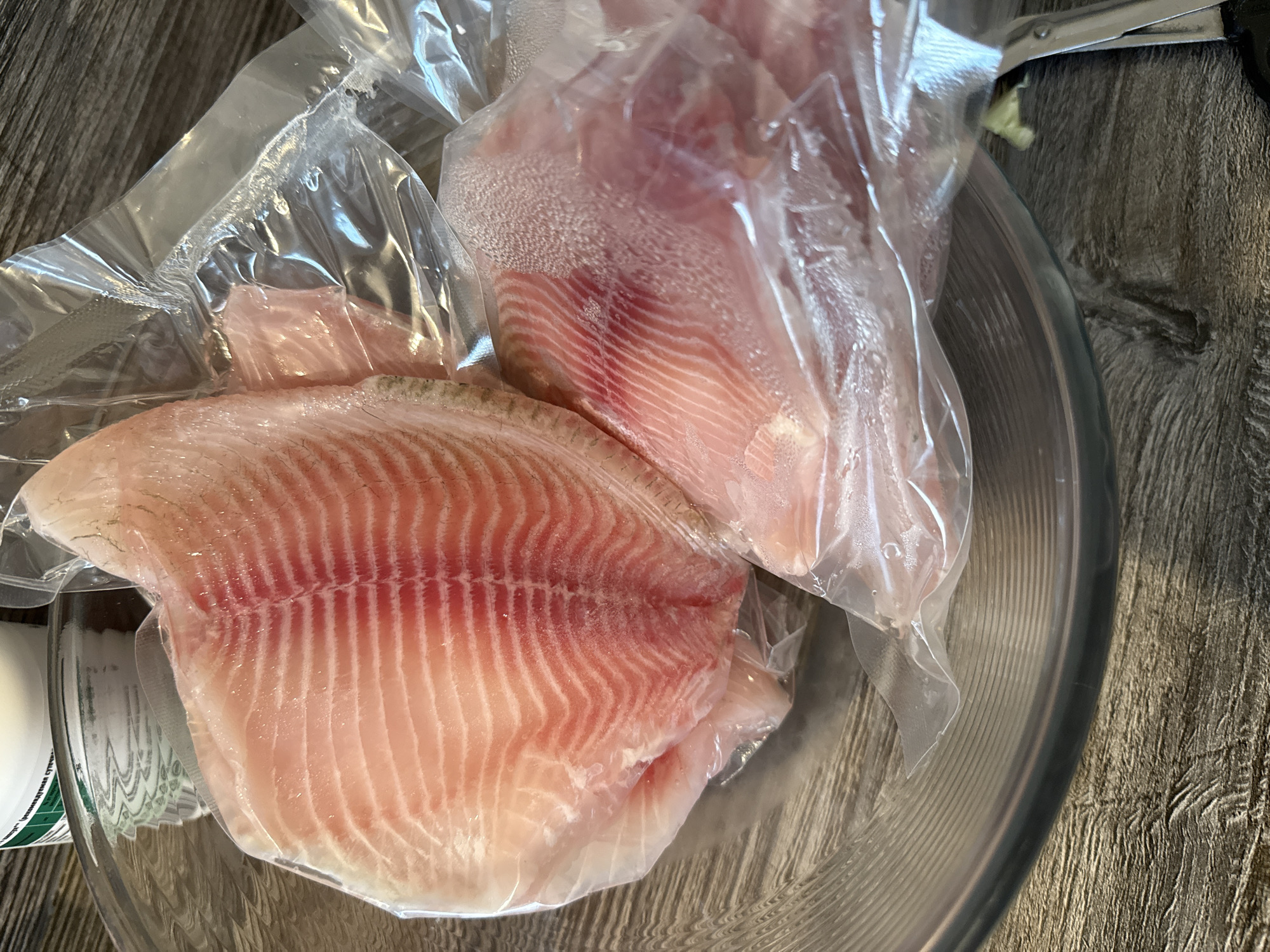 The image shows two vacuum-sealed fish fillets, likely tilapia, within a clear glass bowl. The fillets are pale pink with visible red striations indicating muscle fibers. One fillet is larger and positioned at the top left of the bowl, while a second, smaller fillet is visible beneath it and to the right. The bowl rests on a wooden surface with a visible grain pattern. A portion of a white and green label from a container is also visible in the upper left quadrant of the image. The scene appears to be indoors, likely in a kitchen environment, but no specific location in the city of Perm, Russia can be determined from the visual information provided. There are no people or any actions depicted, only the raw ingredients.