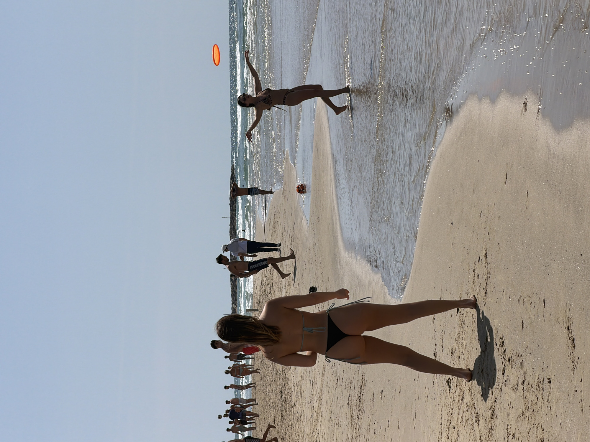 The image depicts a beach scene, likely in Tel Aviv, Israel. The foreground shows a woman in a bikini standing on the sand. Further down the beach, a group of people appear to be engaged in some form of exercise or activity. In the distance, another woman in a bikini is seen playing with a red frisbee. The beach has light-colored sand and the ocean is visible, with waves coming in to shore. The sky is clear and blue.
