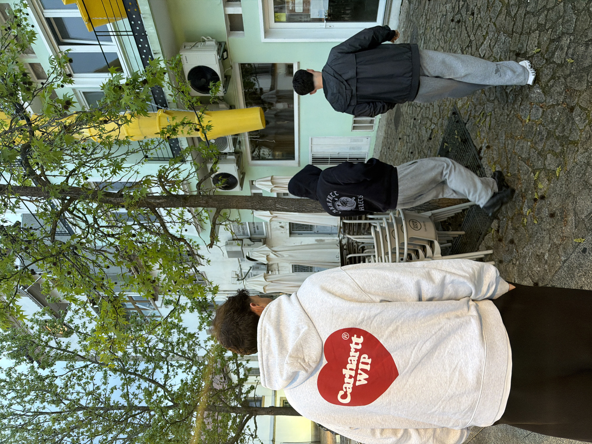 The image captures a scene in Bragança, Portugal. Three individuals are present. The closest person wears a light gray hoodie with a heart-shaped logo containing the words "Carhartt WIP." The second person is dressed in a dark hoodie and light gray sweatpants. The third person is wearing a dark jacket and light gray sweatpants. The setting appears to be outdoors on a paved area. Tables and stacked chairs suggest a cafe or restaurant setting. A building is visible in the background with air conditioning units mounted on its exterior. Yellow piping or ductwork is also attached to the building. Green foliage from a tree is prominent in the foreground, partly obscuring the view of the building.