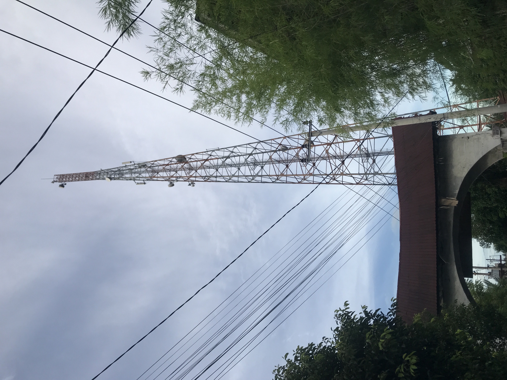 The image shows a tall transmission tower in Lhokseumawe, Indonesia. The tower is constructed of a lattice of metal beams, painted white and red. It is positioned next to a building with a curved roof. Power lines run across the foreground of the sky. A bamboo tree is located in the upper portion of the image. Green bushes are in the bottom corners. The sky is overcast.
