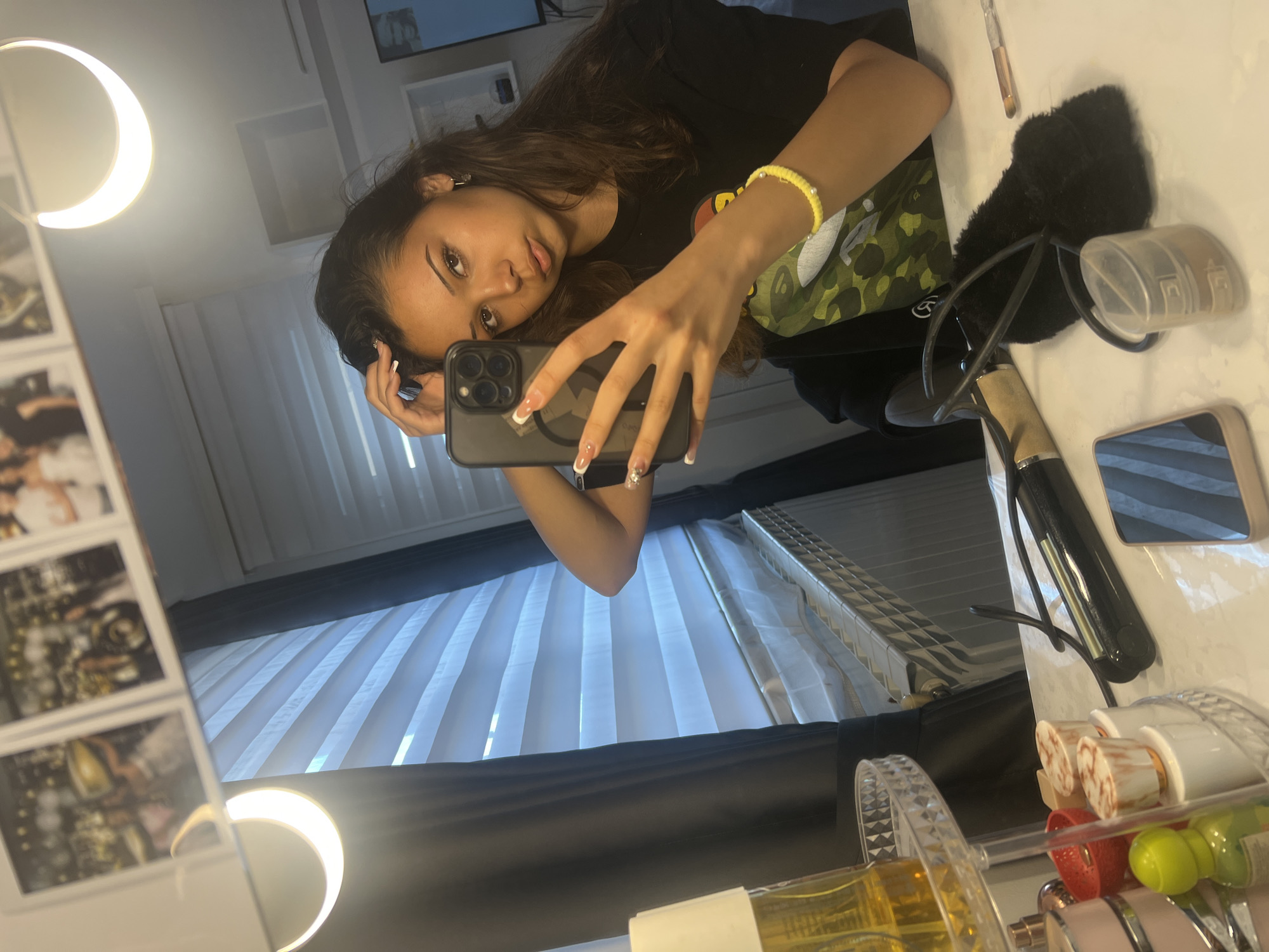 A young woman is taking a selfie in front of a vanity mirror. The reflection shows that she is wearing a black t-shirt, has long brown hair, and is holding a black smartphone. Her nails are manicured with white tips. On the vanity, there are various items including a black fluffy object (possibly a hair accessory), a container, a brush, a black hair straightener, a phone, and a rotating cosmetic organizer filled with items. The background reflected in the mirror includes a window with white blinds. There is also a crescent-shaped light attached to the mirror. The location is Unknown, Unknown.