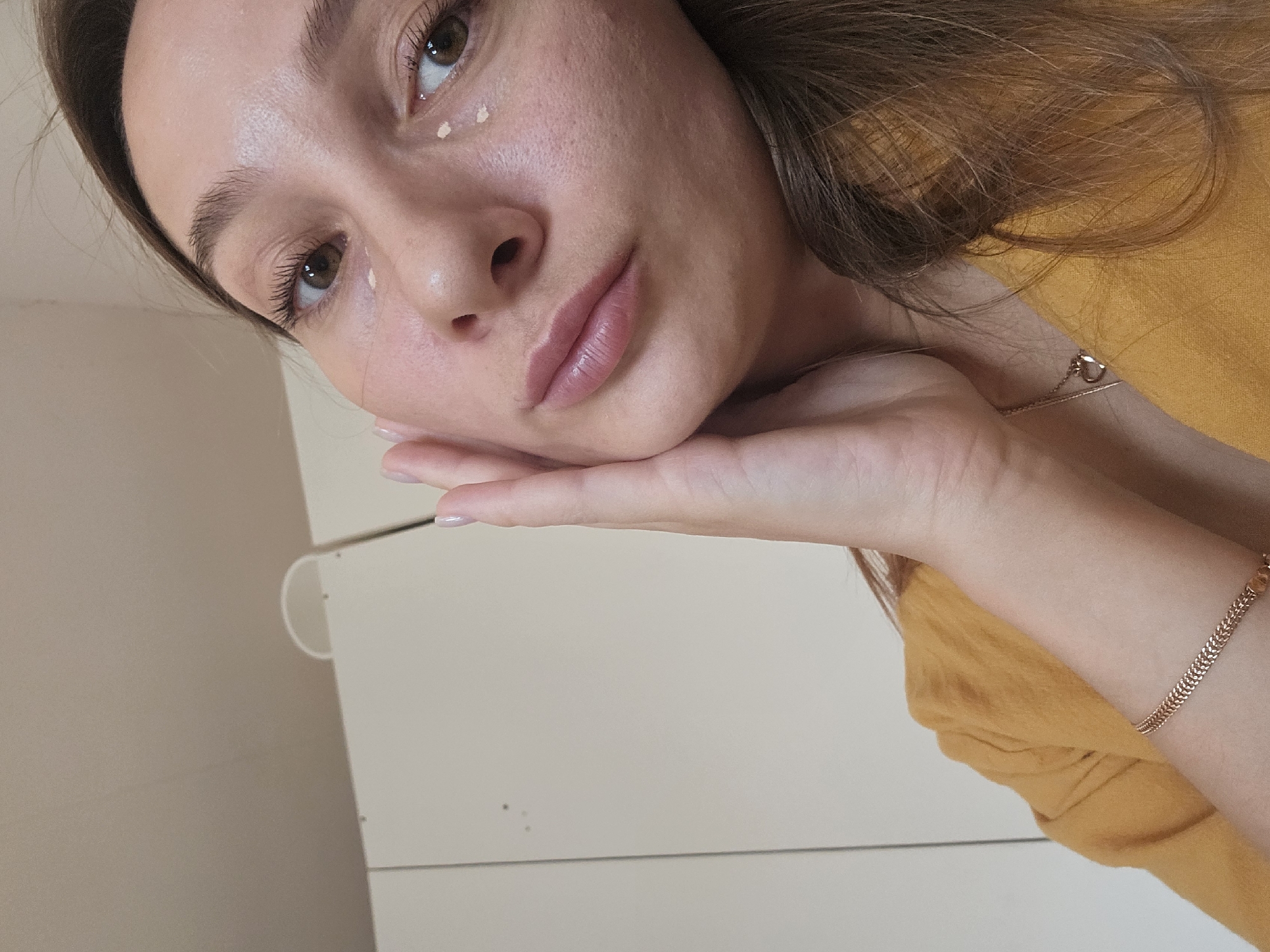 A female individual is depicted in a close-up, head-and-shoulders shot, identified as being in the city of Chaikovsky, Russia. She has brown eyes and long brown hair that cascades over her right shoulder. Her right hand, displaying manicured nails with light pink polish, rests against her right cheek and jawline. Several small, light-colored dots, consistent with a cosmetic product, are visible under her eyes: two beneath her right eye and one beneath her left eye. She is wearing a mustard yellow top, a delicate gold-colored chain necklace with a heart-shaped pendant, and a gold-colored chain bracelet on her left wrist. The background consists of light-colored, possibly off-white, modular cabinet doors with vertical seams. A partial white, D-shaped handle is visible atop one cabinet section. The wall above the cabinets is a plain, light beige. The scene is indoors with soft, ambient lighting.