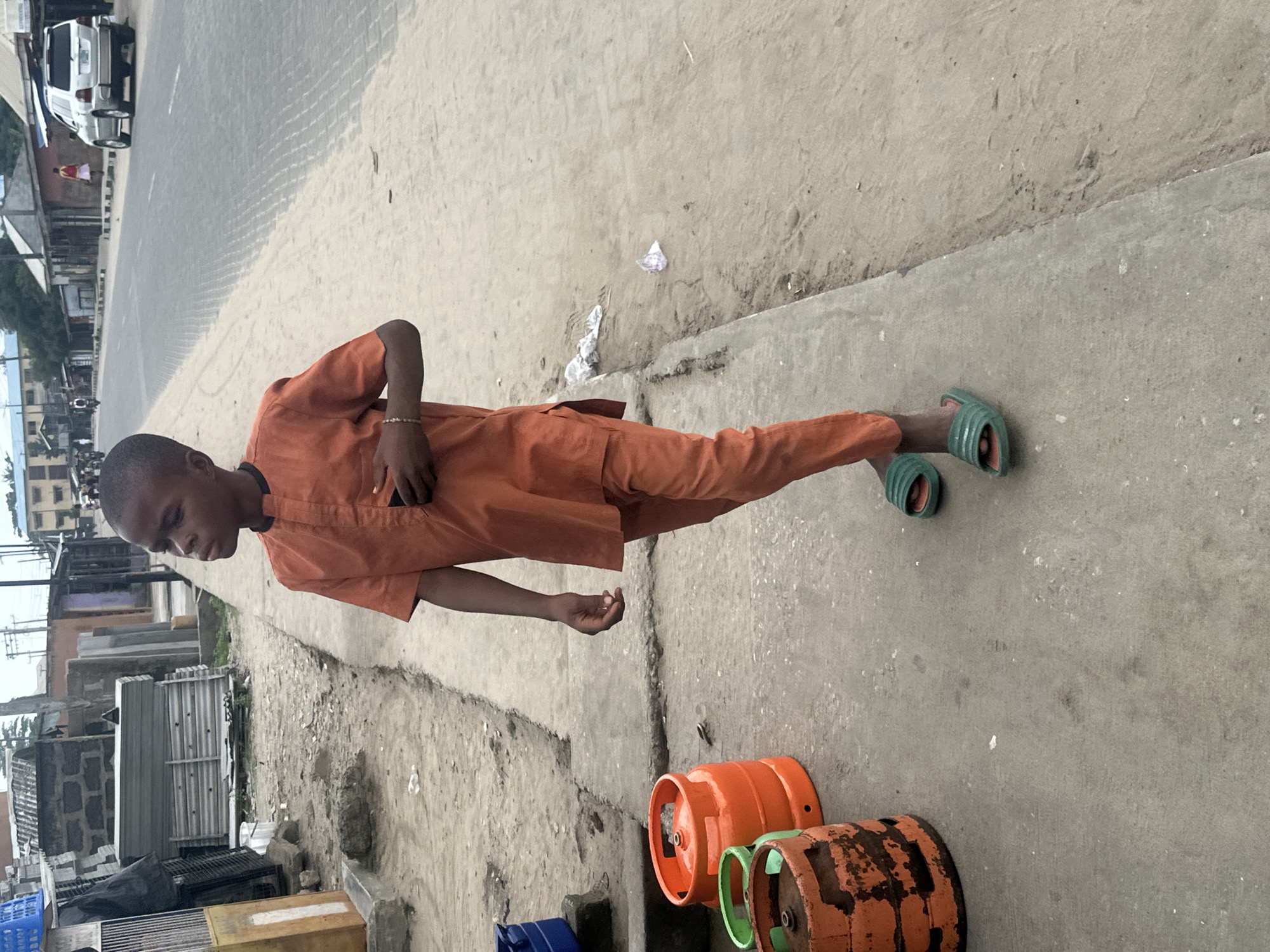 A young boy in an orange outfit and green slides stands on a concrete sidewalk in what appears to be a commercial area of Badagry, Nigeria. To his left, three gas cylinders are visible, two orange and one green. The boy is looking to his right, with his right hand inside his pocket. In the background, a paved road stretches into the distance, with various buildings lining the sides. A silver SUV is parked on the right side of the road. The ground is a mix of sand and concrete, with some debris scattered around. The overall impression is one of a typical street scene in a Nigerian town.