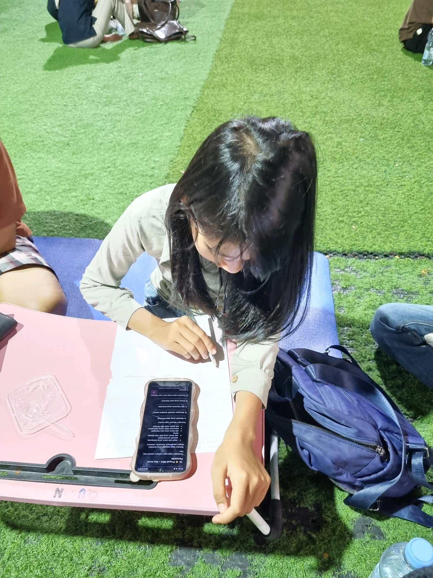In Tanamodindi, Indonesia, an overhead shot shows a young female student, with long dark hair obscuring her face, engrossed in her studies on a bright pink lap desk. She wears a light-colored long-sleeved shirt. A blue backpack rests to her right on the artificial turf. A smartphone displaying text is positioned on the lap desk, next to a pen in her hand, indicating she is actively engaged in writing or reviewing notes. The background features green artificial turf, suggesting an outdoor or recreational study setting. Other individuals are visible in the background, one seated with a dark bag, and another in denim jeans, their presence contributing to the communal atmosphere of the location. A plastic bottle of water is partially visible in the lower right corner of the frame.