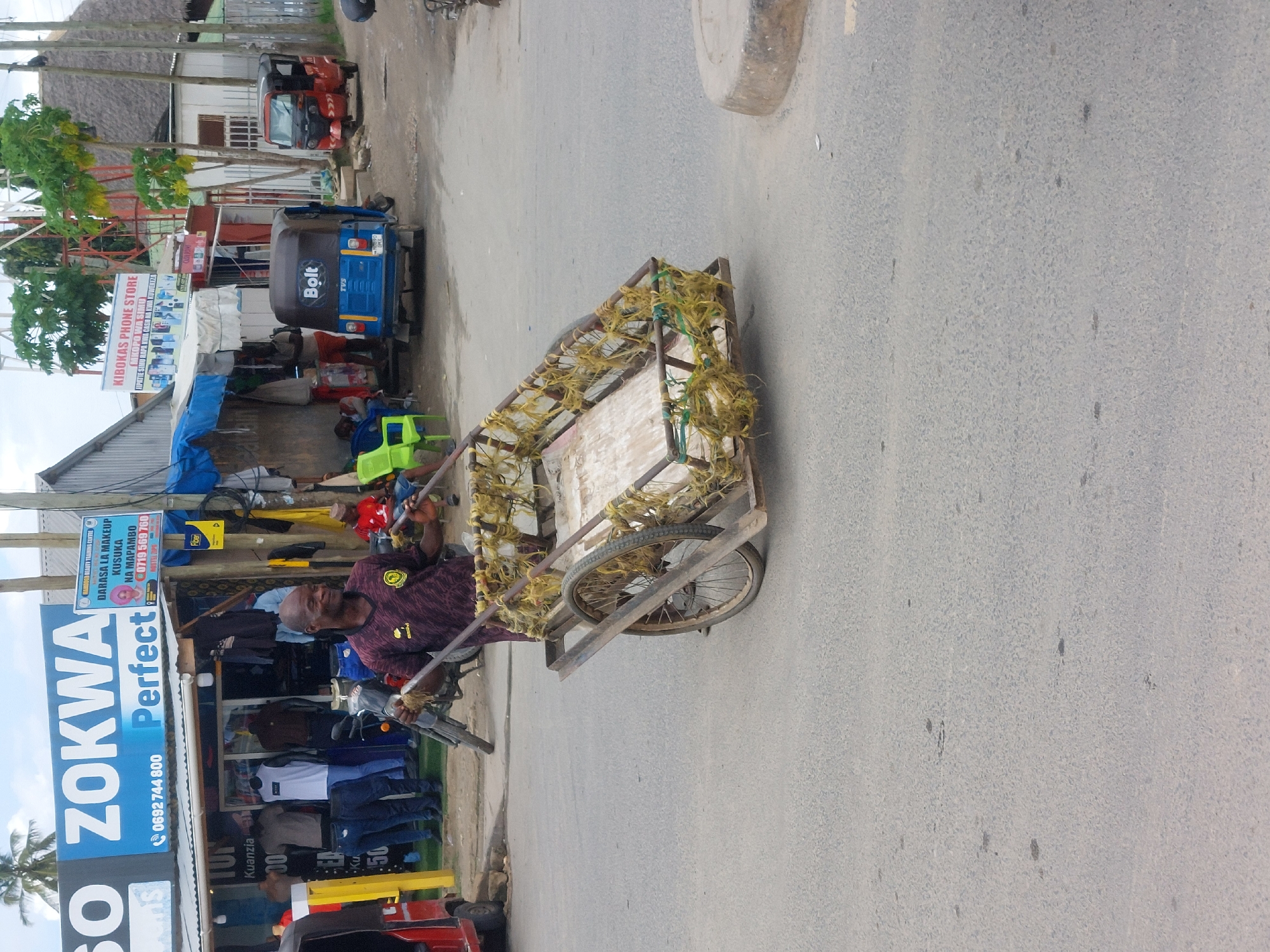 A man is pulling a cart with two bicycle wheels down a street next to a building in Dar es-Salaam, Tanzania. The building has a sign that reads "ZOKWA Perfect" and "KIBOKAS PHONE STORE." Parked behind the cart puller are a bolt and two vehicles. The cart is decorated with strings.