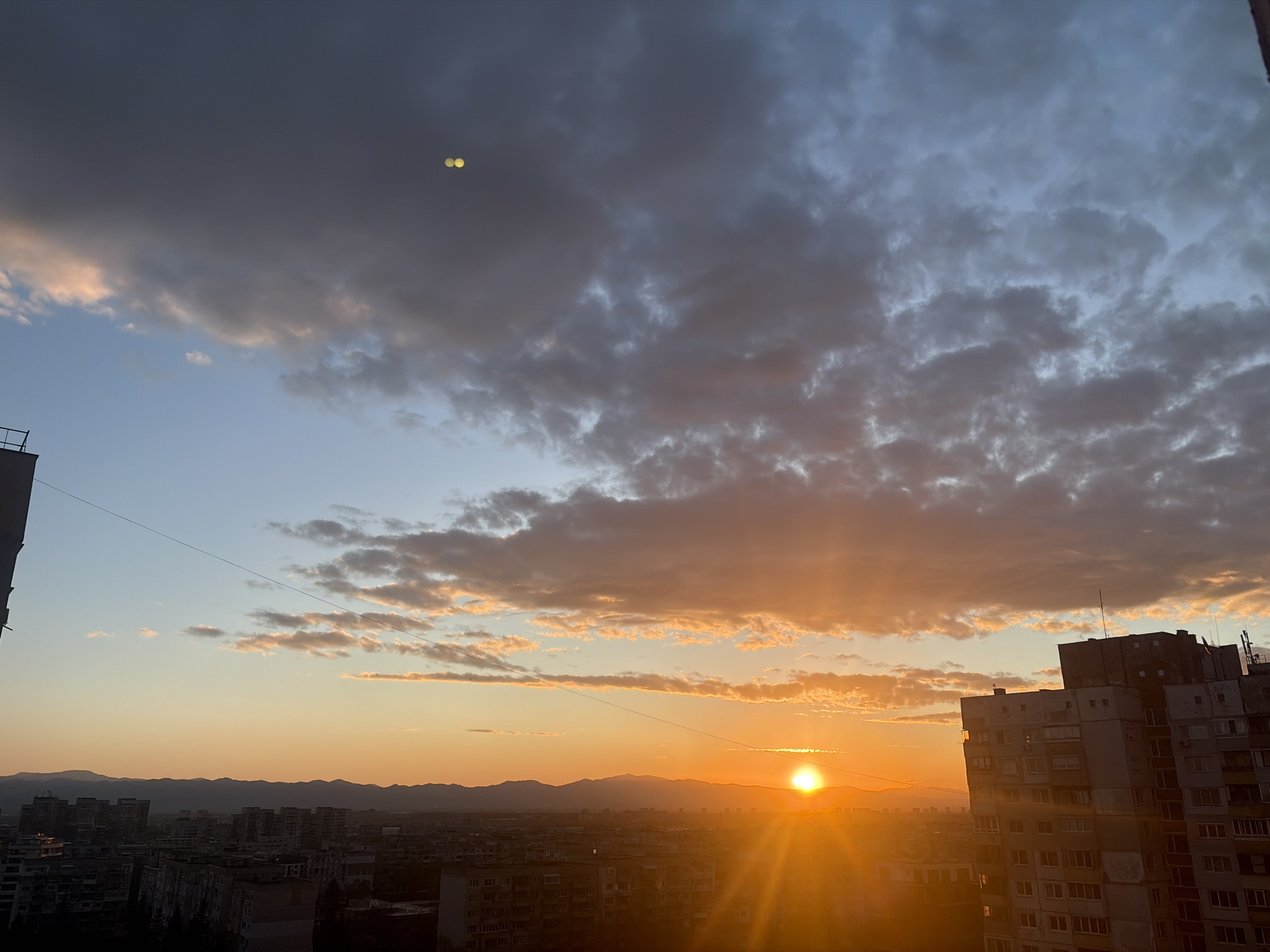 The image shows a sunset over the city of Sofia, Bulgaria. The sun is setting behind distant mountains, with visible sun rays. In the foreground, a tall residential building is on the right, and other buildings are spread across the cityscape. The sky is a mix of blue and gray clouds, with some clouds reflecting the orange light of the setting sun. There is a thin wire going across the middle of the image.
