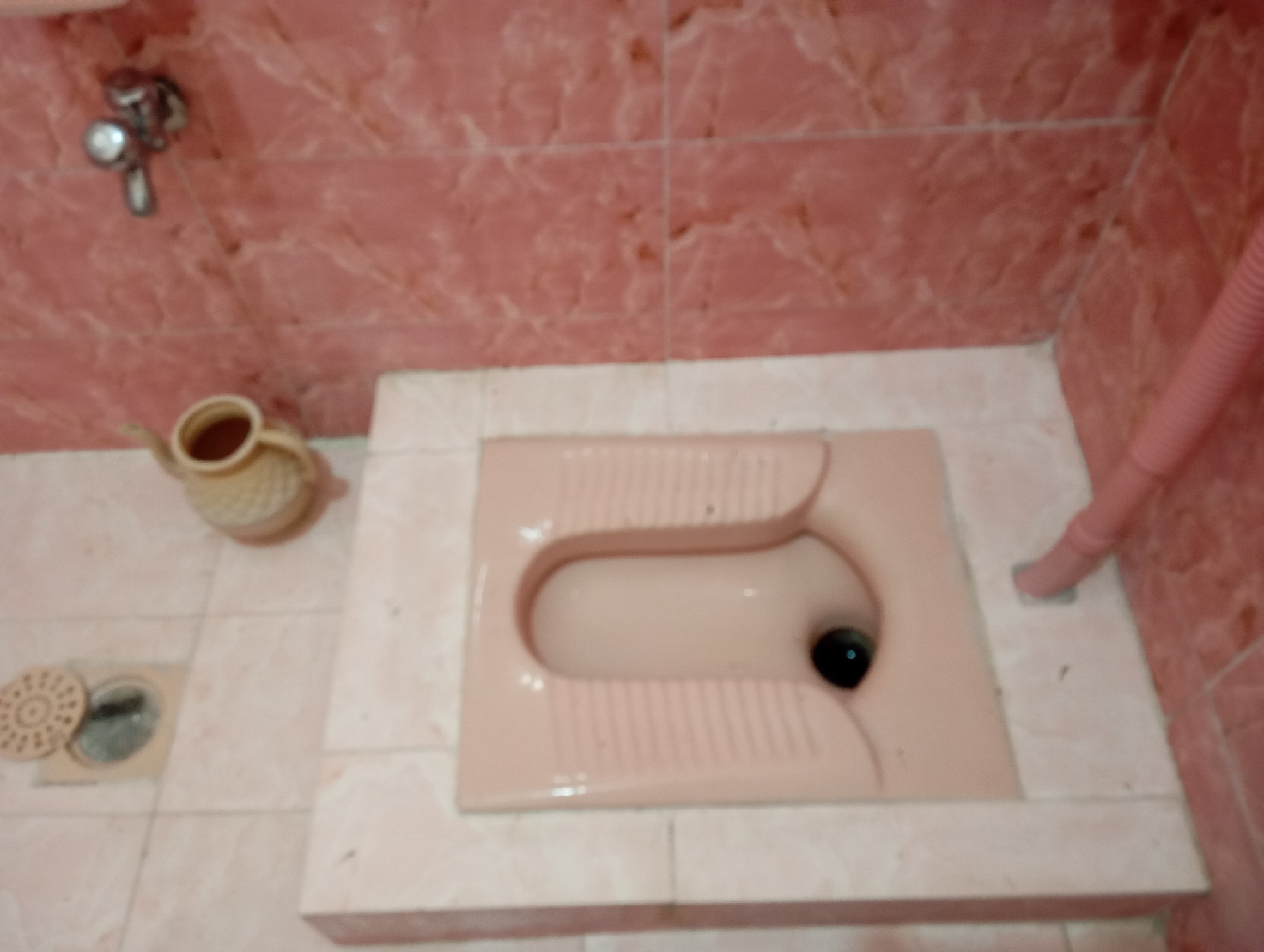 The image shows a squat toilet in a bathroom. The toilet is light pink and appears to be made of ceramic. It is a Western-style squat toilet with a large, oval opening. There is a faucet on the wall above the toilet, and a pink corrugated pipe is visible on the right side of the image. The floor is tiled with light-colored tiles, and the walls are tiled with pink tiles. There is a small pitcher on the floor to the left of the toilet, and a drain cover is also visible on the floor. The image is blurry and out of focus, making it difficult to discern fine details. The scene is likely in a residential bathroom in Dangari, Pakistan.