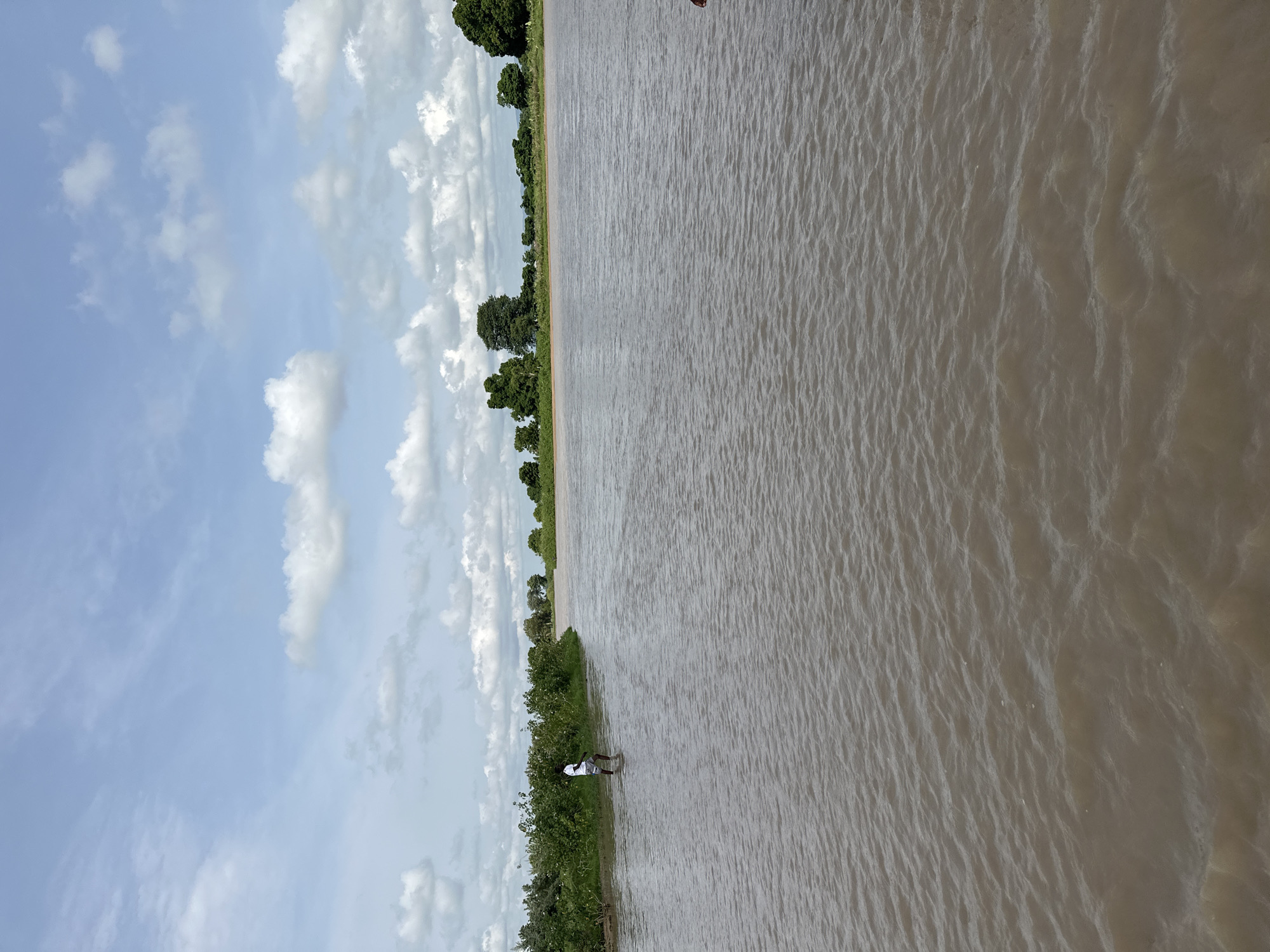 A wide, muddy river flows under a partly cloudy sky in Kunini, Nigeria. On the left bank, a person wearing a white t-shirt and shorts stands in the shallow water near lush green vegetation. The river extends to the horizon, where more trees and vegetation can be seen. The water has ripples on its surface, suggesting a gentle current.