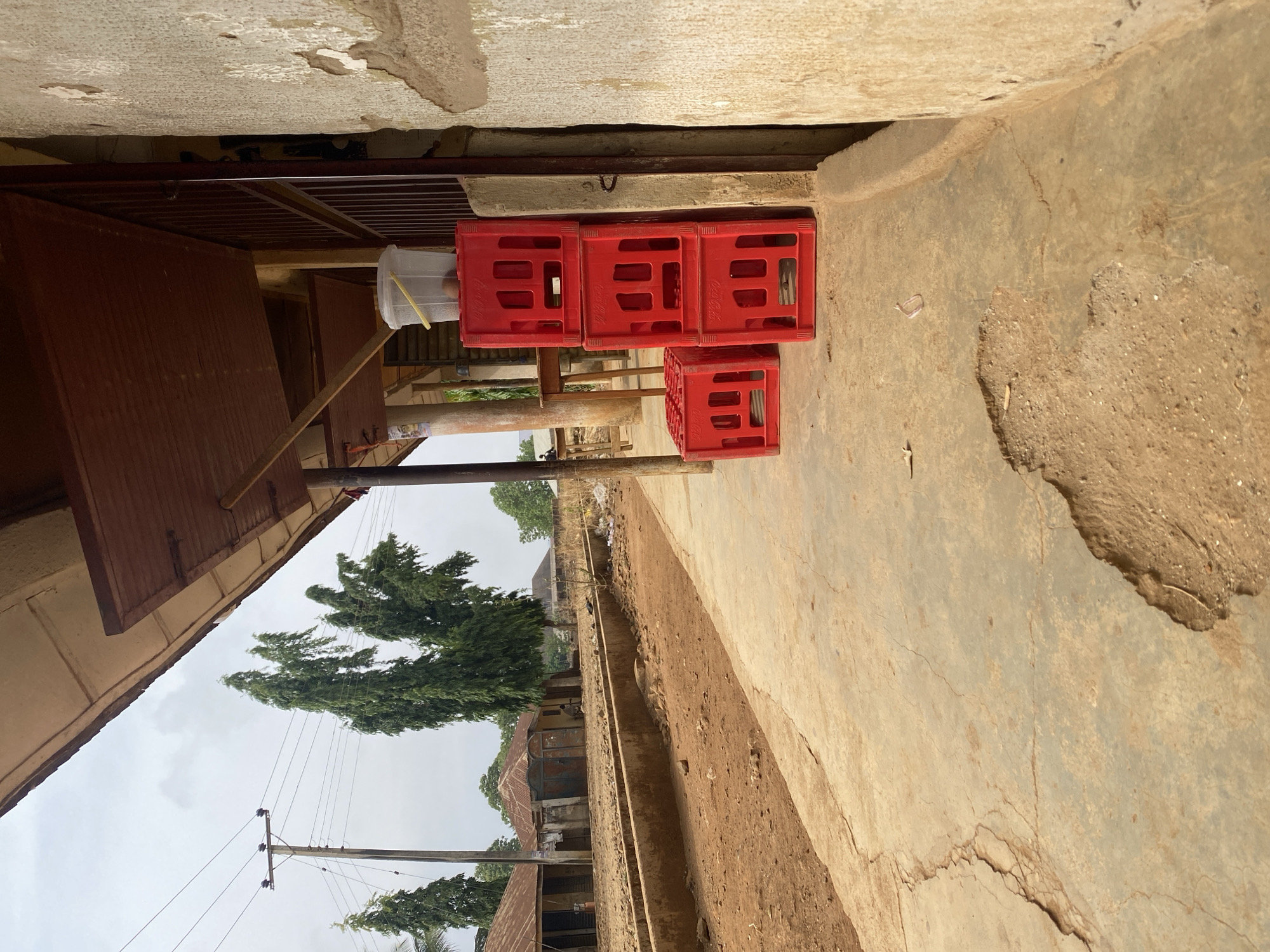 The image shows an outdoor scene in Maigizo, Nigeria. Three red Coca-Cola crates are stacked against a wall. Adjacent to them is a makeshift handwashing station with a plastic container and a faucet mechanism. Beyond this immediate area, the background reveals a street or alley with structures, trees, and utility poles visible in the distance. The ground and walls appear weathered, and the sky is overcast.