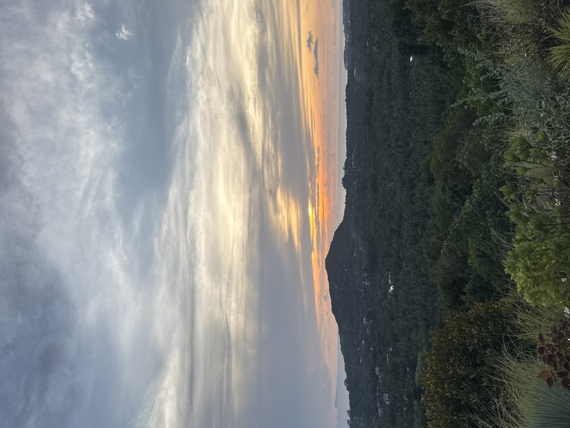 The image depicts a sunset over a lush, forested area. The sky is filled with striated clouds, ranging from pale gray to orange and yellow near the horizon. The forest appears dense and green, with some trees visible on a hillside. There are buildings and small structures nestled among the trees. The location is in the city of Baan Nai Suan, Thailand.