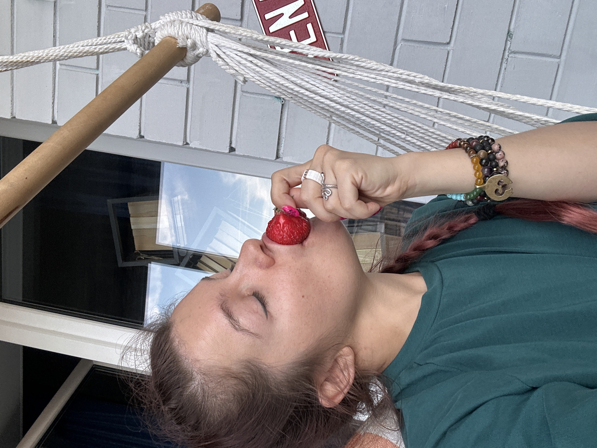 A person is depicted lying horizontally in a white rope hammock, with their eyes closed, taking a bite of a fresh red strawberry. The subject appears to be enjoying a moment of relaxation. Their right hand, holding the strawberry, features a silver-colored ring with a snake design on one finger and a patterned white ring on another. Their fingernails are painted pink. On their right wrist, multiple beaded bracelets are visible, showcasing a variety of colors including dark, light, and a golden charm resembling an 'Om' symbol.
The person is wearing a dark green t-shirt, and their hair, which has a reddish hue, is partially braided and styled over their shoulder. The hammock's wooden spreader bar is visible on the left side of the frame. In the background, there is a light-colored, possibly white or grey, tiled or brick wall. Below the hammock structure, a window reflects a bright, cloudy sky, suggesting it is daytime. A red sign with white lettering, partially visible as "EN", is mounted on the wall in the upper background. The scene suggests a calm, leisure activity taking place in Vladimir, Russia.