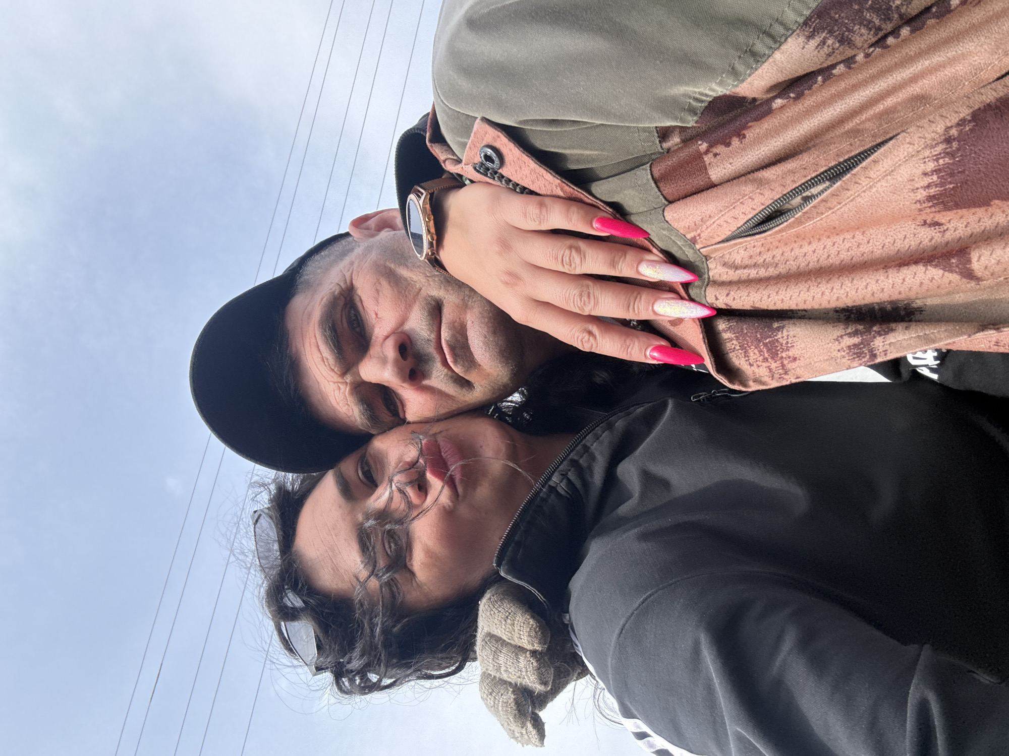 A man wearing a dark-colored baseball cap and a woman wearing sunglasses, a dark jacket, and gloves are posing for a selfie outdoors. The background shows overhead power lines against a cloudy sky. The man appears to be wearing a watch and a jacket that is green and a light pinkish-brown color. The woman is kissing him. A hand with long, artificial nails is visible in the upper-right of the frame. The location is outdoors, potentially near power lines. The exact location in the city of Кемуль, Russia cannot be determined from the image.