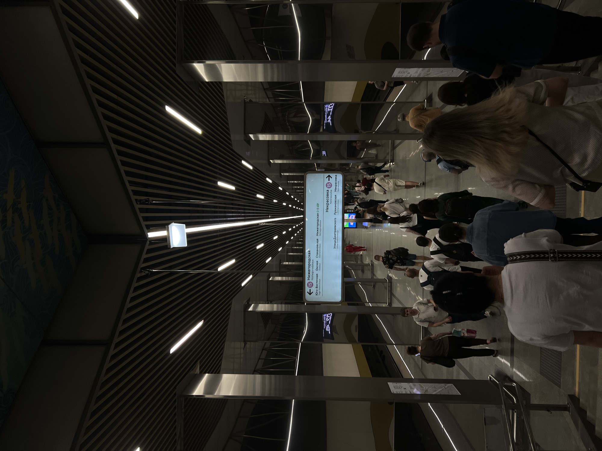 Here is a description of the image:
The image shows a subway station in Moscow, Russia. The station is modern, with a sleek design. There are a lot of people walking in the station. The station sign indicates directions for different train lines and stations, some of which are "Nizhegorodskaya," "Nekrasovka," "Stakhanovskaya," "Okakaya," "Nichegors," "Ulitsa Dmitriyevskogo," and "Lukhmanovskaya." The walls are decorated with horizontal stripes and illuminated by long, linear lights.