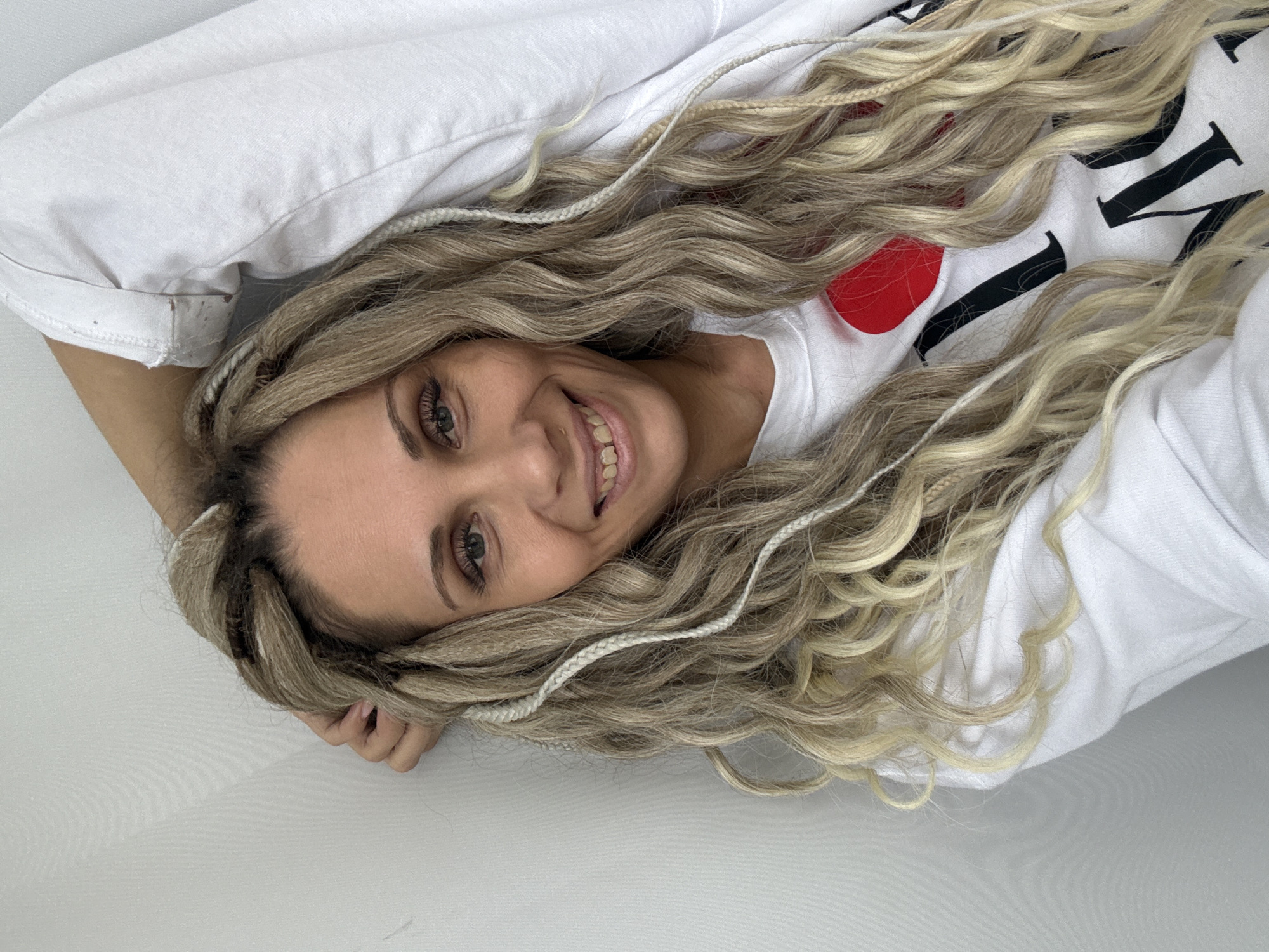 A woman with braided blonde hair lies on a white surface. She is wearing a white t-shirt with a red heart graphic and black lettering. Her head is tilted, and she is smiling while looking directly at the camera. One hand rests near her head. The background is a plain white surface. The location is unspecified, and no information indicates it is Rusanovo, Russia.