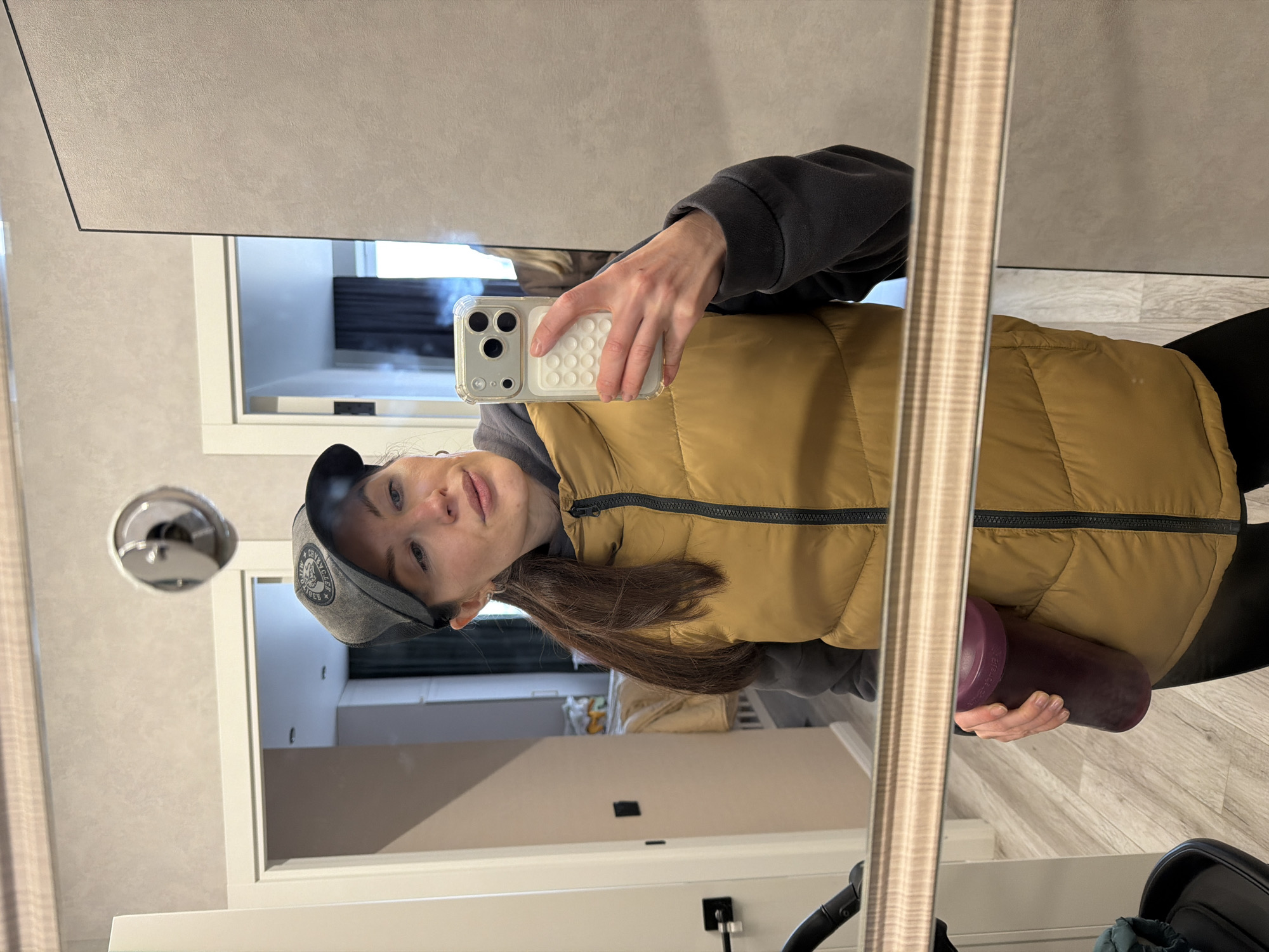 A young woman is depicted taking a mirror selfie in an indoor setting in Perm, Russia. She holds a smartphone with a clear case and a white pop socket or grip in her right hand, angling it to capture her reflection. In her left hand, she holds a purple bottle, possibly a shaker or water bottle.
She is dressed in casual attire, including a dark grey long-sleeved top, a mustard yellow puffer vest with a black zipper, and black leggings. She wears a grey baseball cap with a dark brim, featuring a partially visible white logo or text that appears to be "CRRYBLCK" or "CRYBLK." Her long, dark brown hair cascades over her shoulder.
The background reflected in the mirror shows a clean, modern interior, likely a hallway or room in an apartment or hotel. Visible elements include white doorframes, light-colored walls, and a light wood-patterned floor. A circular, reflective light fixture is visible on the ceiling in the reflection. The scene is well-lit, suggesting daytime. The woman has a neutral expression, gazing towards the camera.