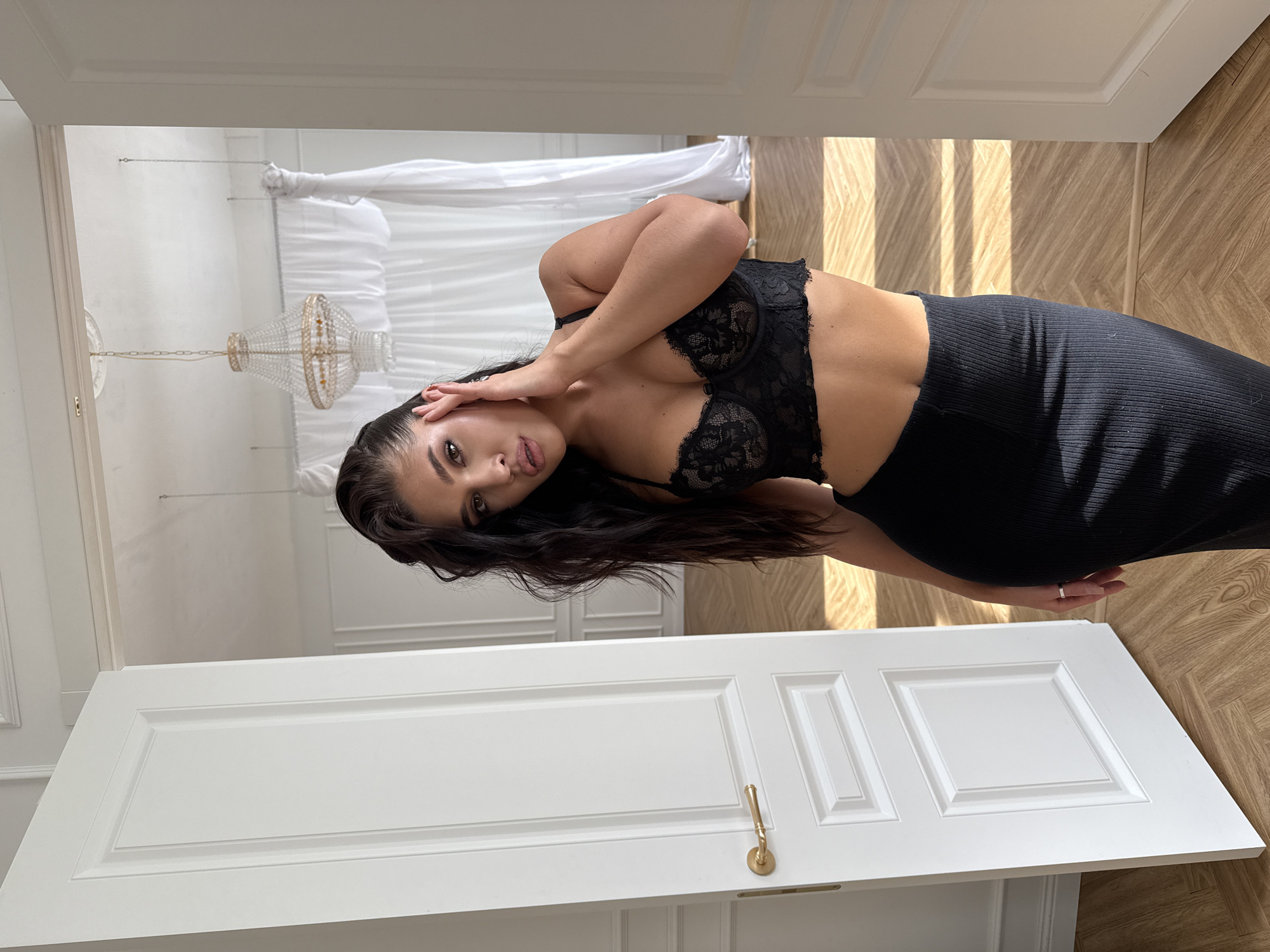 A woman poses in a room. She is wearing a black lace bra and a black ribbed skirt. Her hair is long and dark. She is standing near a white door with a gold handle. Behind her, there is a chandelier and white curtains. The floor is wooden. The location is Kazan, Russia.