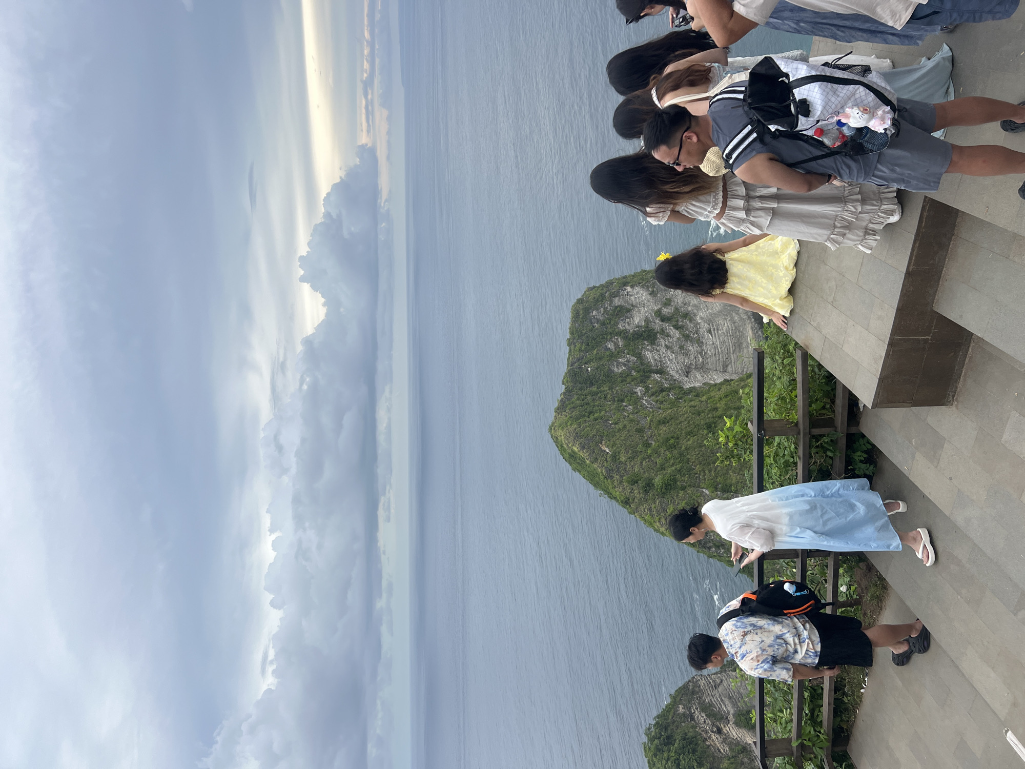 The image captures a scenic view from a cliffside viewpoint in Nusa Penida, Indonesia. The perspective is from a paved platform where several people are gathered, looking out over a vast expanse of ocean.
In the foreground, a group of people stands along a wooden railing. Most are seen from behind or in profile, focusing their attention on the panoramic natural beauty. Among them is a man in a grey striped t-shirt and dark shorts, wearing glasses and carrying a white and black backpack adorned with small items including water bottles and a plush toy. Next to him, a woman in a light beige, ruffled dress also gazes at the view. A small child in a bright yellow dress is also present, looking towards the ocean. Further along the railing, another couple is visible; one person wears a light blue and white flowy dress, and another is in a patterned shirt and black shorts with a black and orange backpack.
Beyond the viewing platform, a dramatic landscape unfolds. A deep blue-grey ocean stretches across the middle of the frame, meeting a cloudy sky overhead. Lush, green-covered cliffs rise steeply from the water, with one prominent, rugged landmass in the mid-ground. The sky is largely overcast, with dense clouds creating diffused lighting, though some brighter patches suggest the sun might be breaking through elsewhere.
The overall activity depicted is people enjoying and observing a breathtaking coastal vista. The scene evokes a sense of awe and appreciation for nature's grandeur. No visible text is present in the image. The time of day appears to be either an overcast midday or a late afternoon with soft, filtered light.