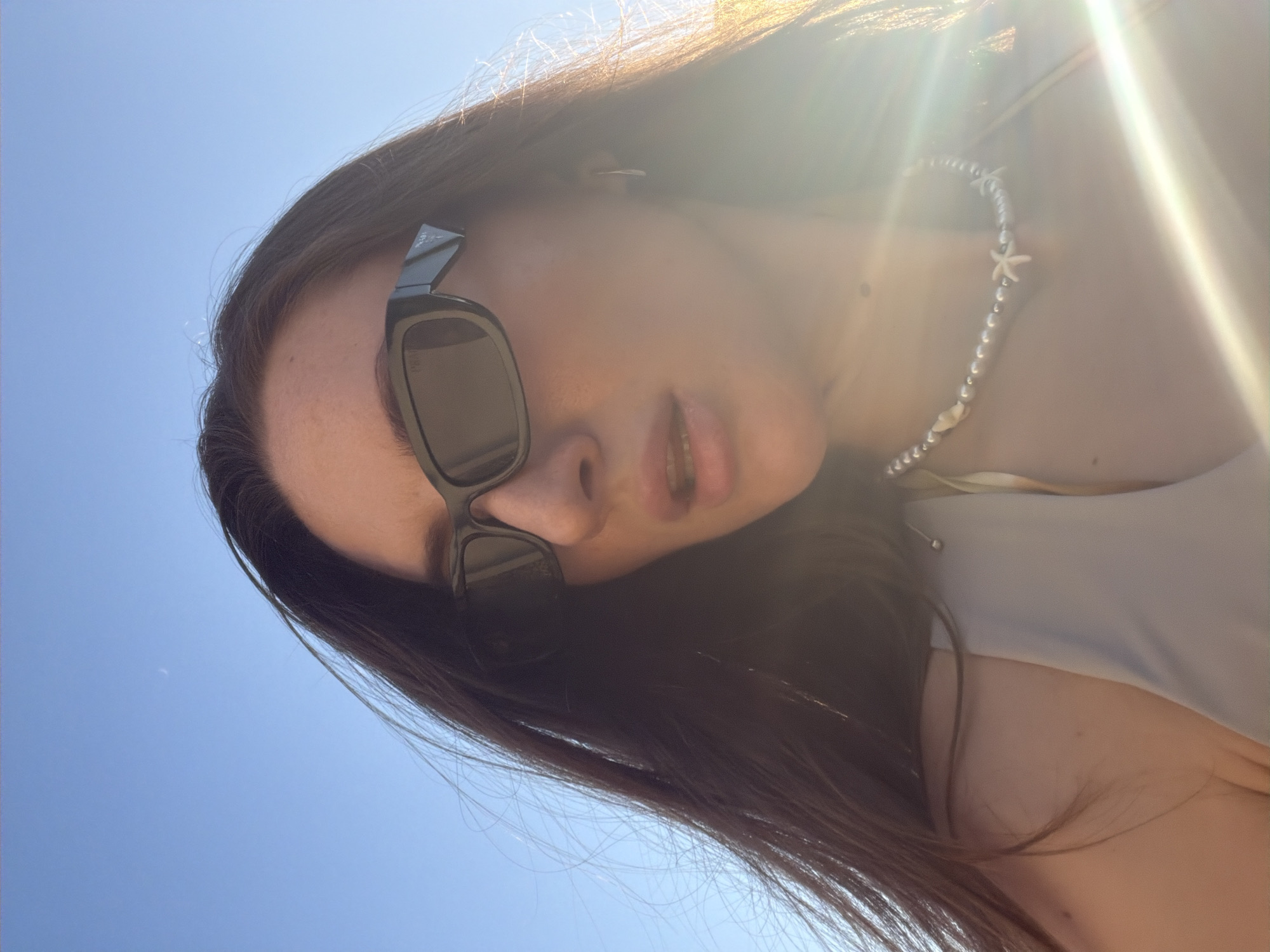 A close-up, low-angle outdoor shot captures a young woman wearing sunglasses and a pearl necklace with starfish charms against a clear blue sky. The woman has long, dark hair that is partially obscuring her face. She is wearing a light-colored top. The sun is bright, causing lens flare and illuminating the right side of her face and hair. The background is a cloudless sky. The image does not contain any specific landmarks or visual cues that would indicate its location within Sharm El Sheikh, Egypt.