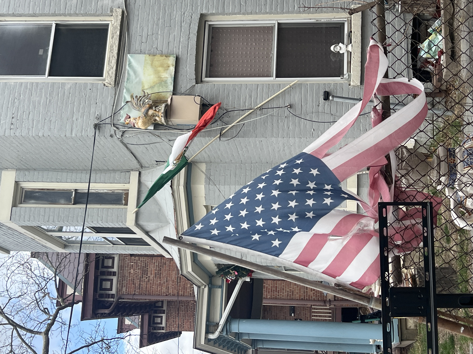 The image captures a scene in what appears to be a residential area, possibly the City of Newburgh, United States of America. The backdrop consists of multi-story buildings with varying architectural styles, constructed primarily of brick and grey siding. Dominating the foreground is a tattered American flag hanging alongside a Mexican flag on a wooden pole. A chain-link fence is also prominently visible in the scene, adding a layer of visual texture. A Christmas wreath is visible between the buildings. Other details include outdoor lights and wall art, providing clues about the local aesthetic.