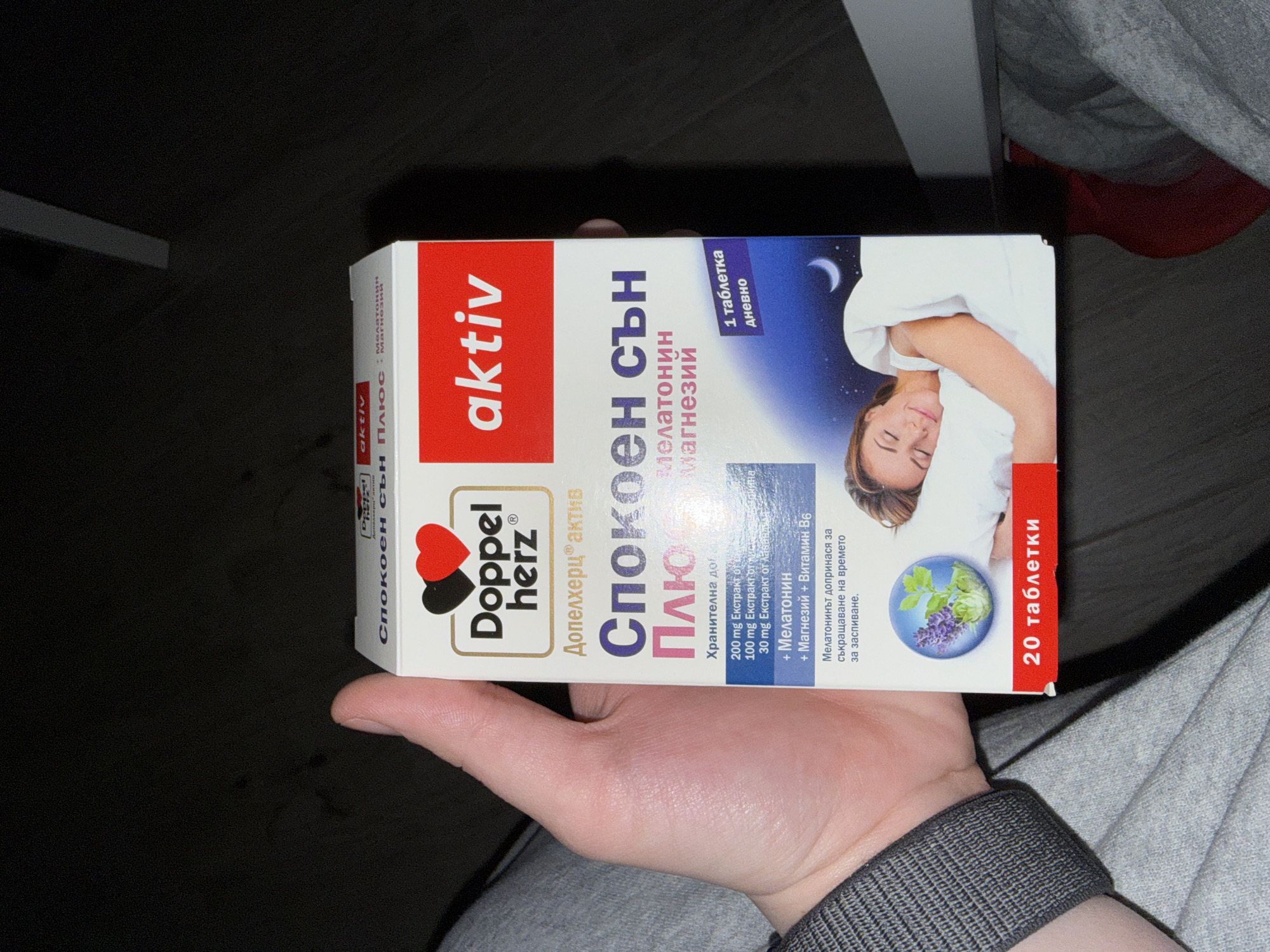 A hand holds a box of "Спокоен сън Плюс" (Relaxing Sleep Plus) dietary supplements by Doppelherz. The box contains 20 tablets and includes melatonin and magnesium. The writing on the box is in Bulgarian. The background is dark and slightly out of focus, showing a table and a grey fabric. I am unable to determine the location to be Sofia, Bulgaria based on the visual information in the image.