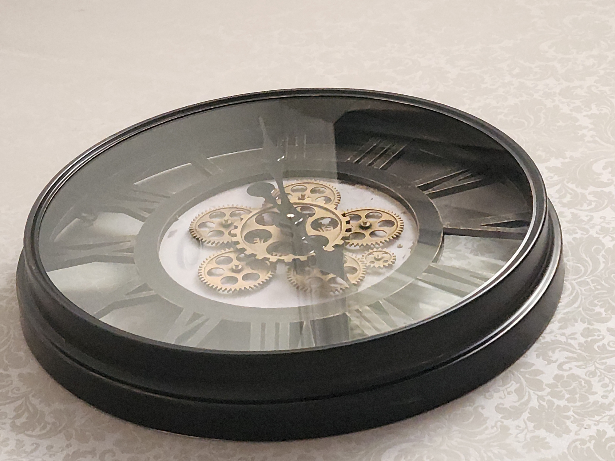 The image shows a close-up, angled view of a black-framed decorative wall clock with Roman numerals. The clock features visible gold-colored gears in its center. The background is a light-colored wallpaper with a subtle damask pattern. The clock is mounted on a wall. The image does not provide any information about the city of Idar-Oberstein, Germany.