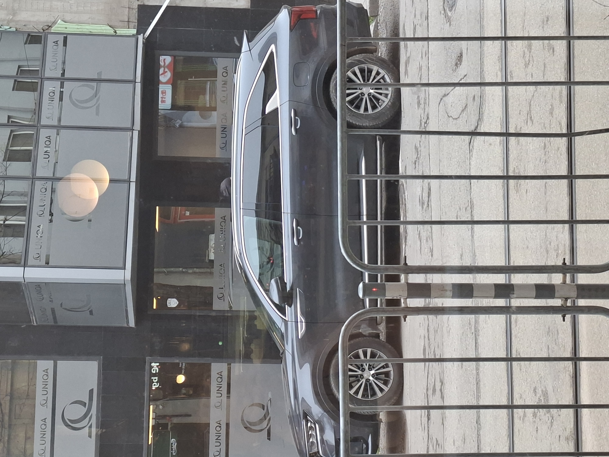 Here's a description of the image:
**Scene:** The image captures a street scene outside what appears to be a UNIQA insurance office in Sofia, Bulgaria.
**Subjects and Objects:**
* A dark-colored SUV is parked next to the UNIQA building.
* A metal fence is in the foreground, partially obstructing the view.
* The building is prominently branded with the "UNIQA" logo on its glass facade. Reflected in the windows are street details, including signage that likely identify the local location.
**Environment:**
* The setting is urban, with a paved street and commercial building facade.