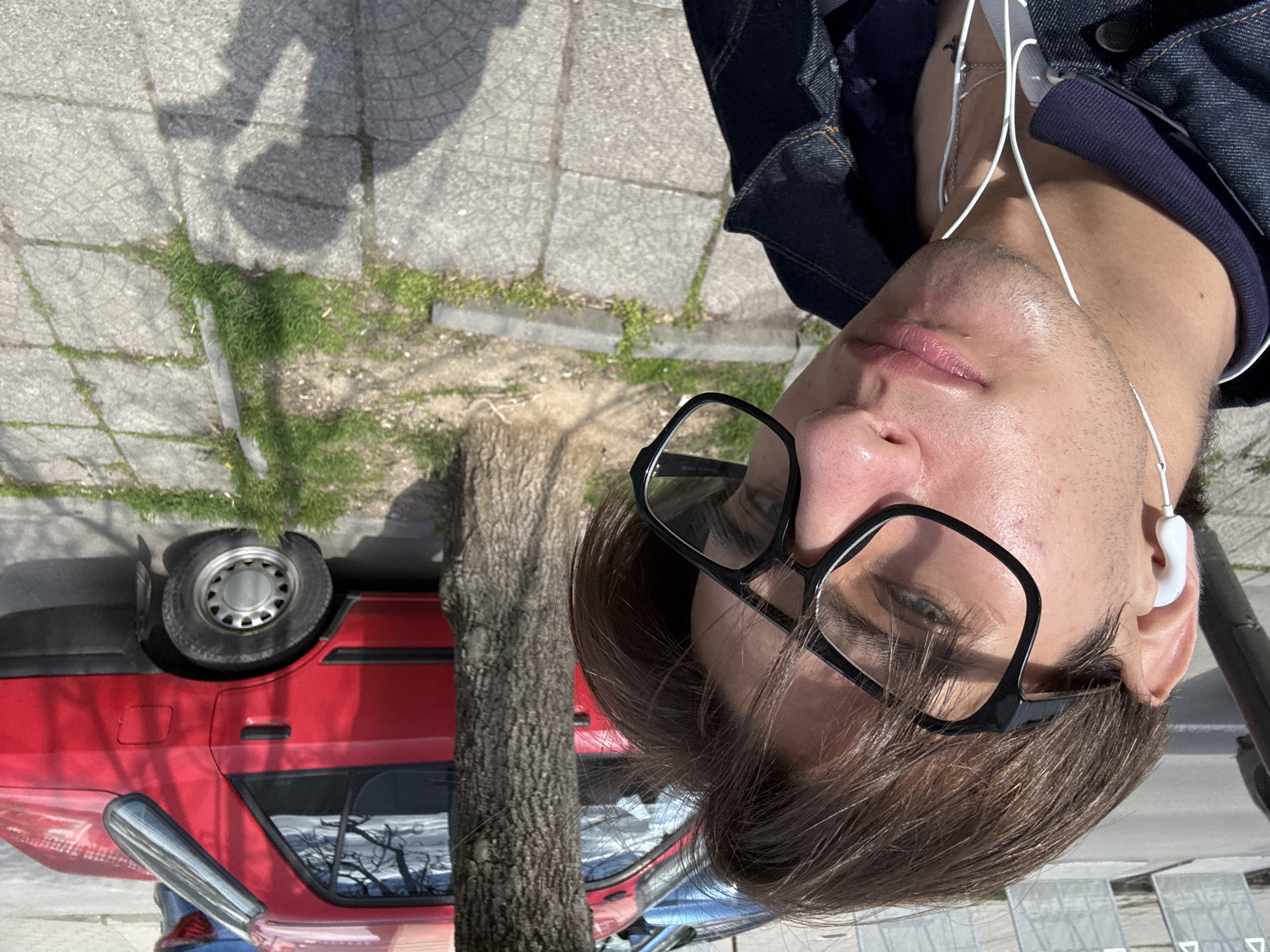 A man is standing on a sidewalk next to a street, wearing glasses and headphones. He is wearing a denim jacket. A red car is parked adjacent to the sidewalk. A tree stands between the man and the car. The setting appears to be urban. While the location cannot be definitively confirmed as Sofia, Bulgaria from visual cues alone, the architectural style, vegetation, and vehicle types are potentially consistent with that region.