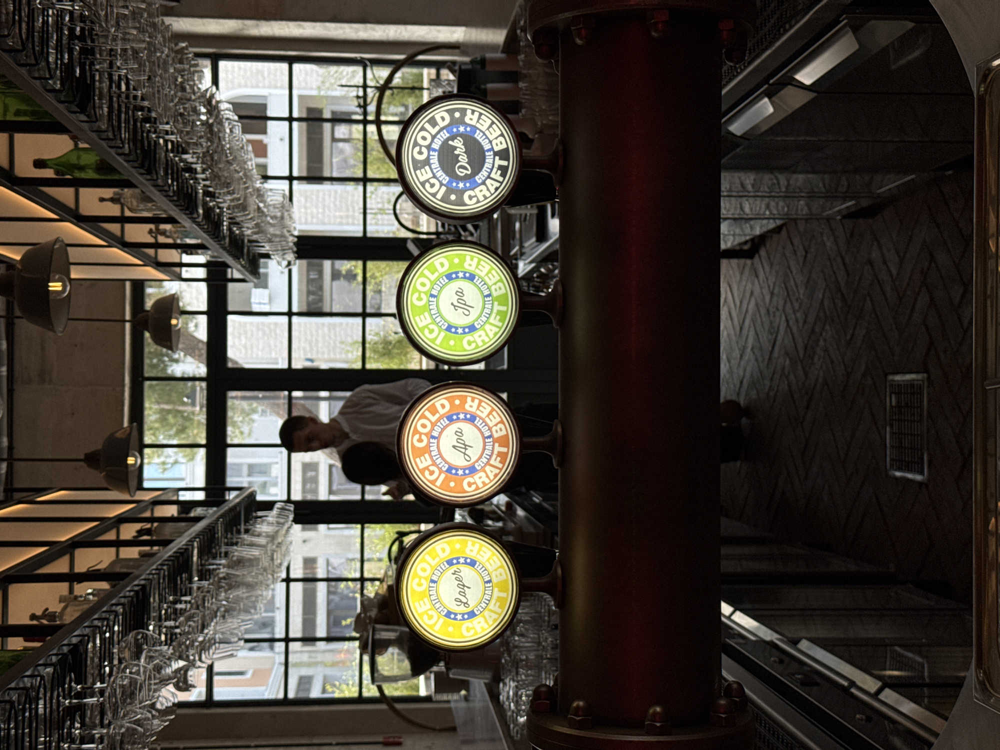 The image displays the interior of a bar or pub, likely located in Radovići, Montenegro, with a focus on its beverage service area. A prominent, dark, pipe-like structure on the right features four illuminated circular beer tap handles, each advertising "ICE COLD CENTRALE HOTEL *** CRAFT BEER." From top to bottom, the specific beer types are "DARK" (blue sign), "IPA" (green sign), "APA" (orange sign), and "LAGER" (yellow sign).
Above the bar, a long, tiered rack is filled with numerous clean, inverted drinking glasses. Through large, grid-patterned windows in the background, an urban outdoor scene with other buildings and hints of greenery is visible, indicating it is daytime. A person, likely a bartender or staff member, is partially seen behind the bar area, looking towards the taps, suggesting the establishment is operational or getting ready for service. Industrial-style pendant lights with exposed bulbs hang near the glass rack. The wall to the far right exhibits a dark, textured herringbone pattern. The "CENTRALE HOTEL" branding on the taps suggests the bar is associated with a hotel.