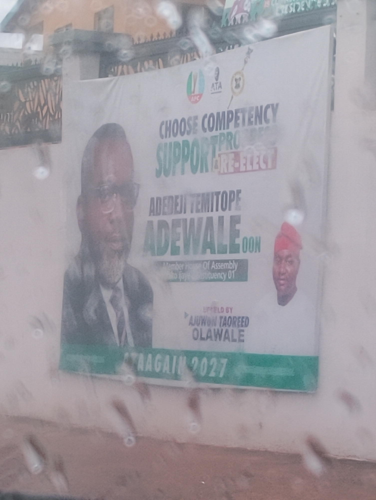 This is a photograph of a political banner displayed outdoors, likely in Iju, Nigeria. The banner features two portraits of men. The man on the left is wearing glasses and a suit, while the man on the right is wearing a red headwrap. The banner has green and white text. Some of the visible text includes "CHOOSE COMPETENCY", "SUPPORT", "RE-ELECT", and names that appear to be "ADEDEJI TEMITOPE ADEWALE" and "AJUWON TAOREED OLAWALE". There is also the year "2027" displayed prominently. The banner itself is white with green and red accents. The background shows a building with decorative fencing. The photograph appears to have been taken through a window or some surface with water droplets on it, creating a slightly blurred and hazy effect. The lighting suggests it is daytime.