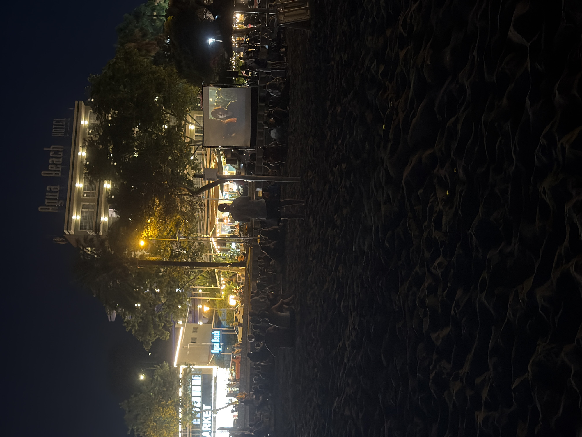 At night on a beach in Calvià, Spain, a group of people is seated on the sand watching an outdoor movie screening. The projector screen is visible in the center of the frame, displaying a movie with a young woman's face illuminated. Behind the screen, buildings with illuminated windows are visible, including a hotel with "Agua Beach HOTEL" signage. Palm trees and other foliage are interspersed between the buildings and the beach. The sand in the foreground is dark and appears undisturbed by the seated audience.