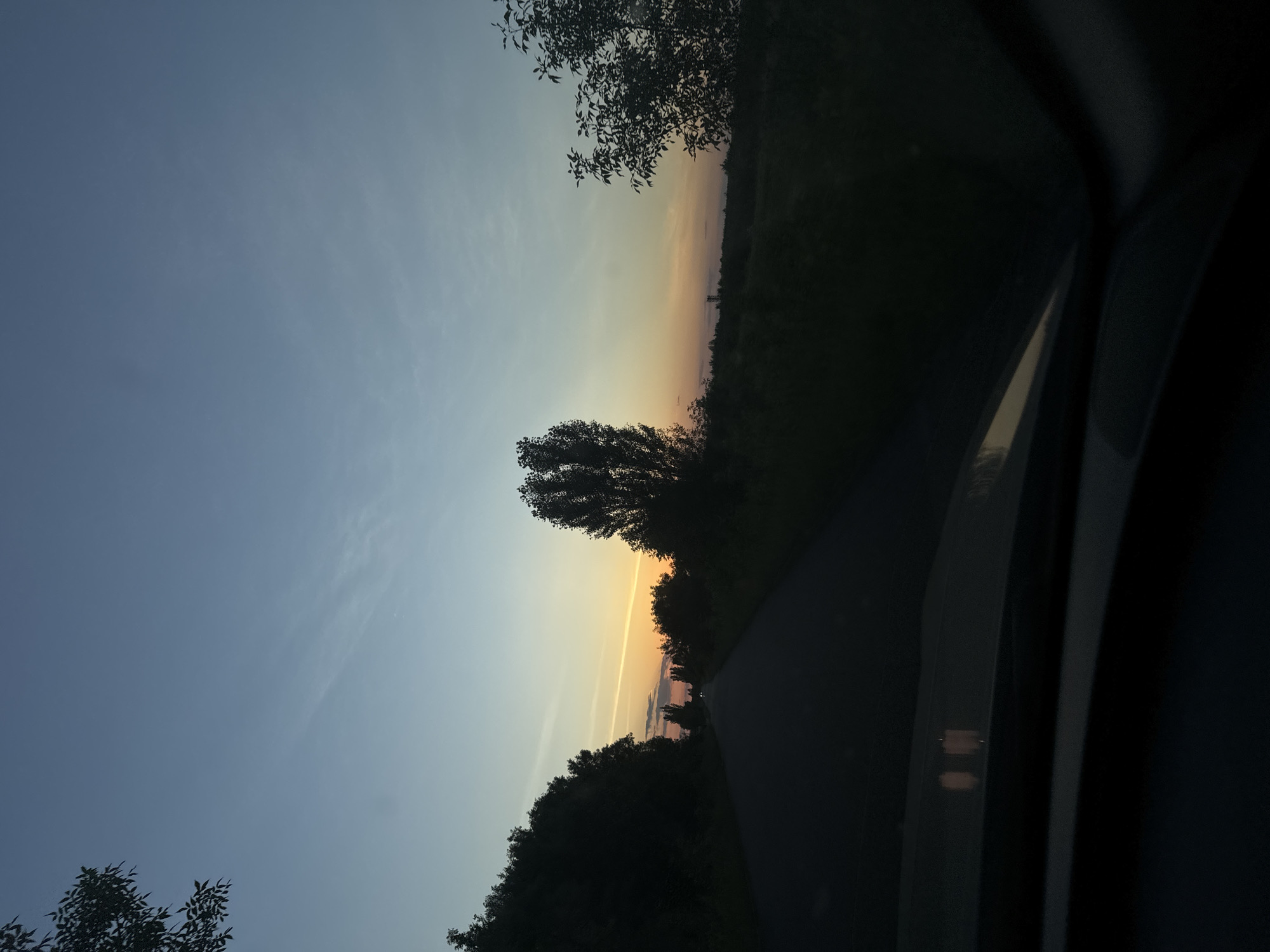 The image captures a serene sunset scene viewed from inside a vehicle driving along a road in the vicinity of Dovhe, Ukraine. The road, paved with asphalt, stretches into the distance, flanked by dense trees on either side. The sky displays a gradient of colors, from a deep blue at the top to warmer hues of orange and yellow near the horizon, indicating the setting sun. A few wispy clouds are visible in the sky. In the foreground, the dashboard of the vehicle is partially visible, with some interior lights illuminated. The overall atmosphere is peaceful and evokes a sense of journey during twilight.
