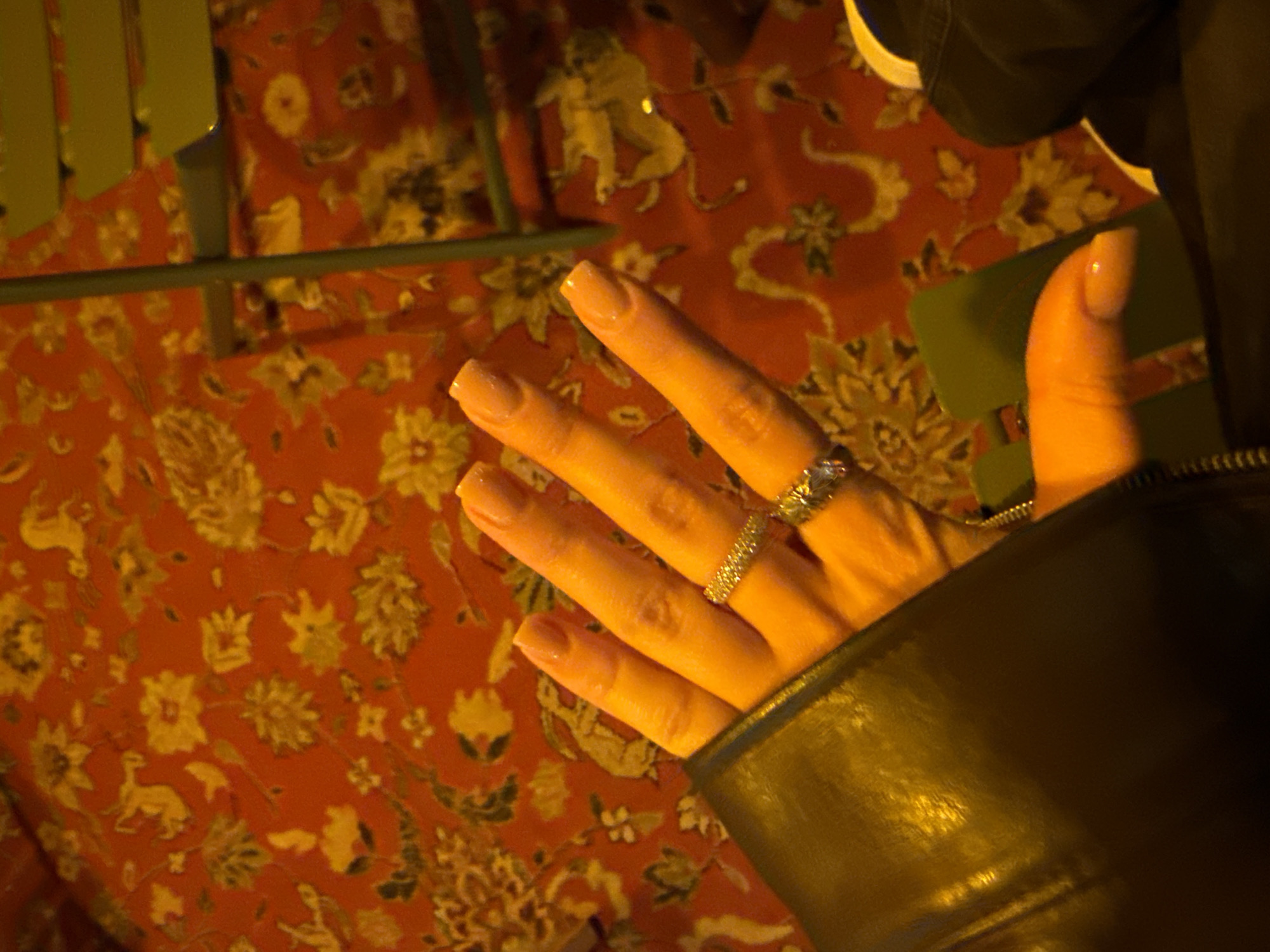 A hand, adorned with two rings, is positioned over an orange patterned tablecloth. A black leather jacket cuff frames part of the hand. The background includes what appears to be green metal furniture. The location is Yerevan, Armenia.
