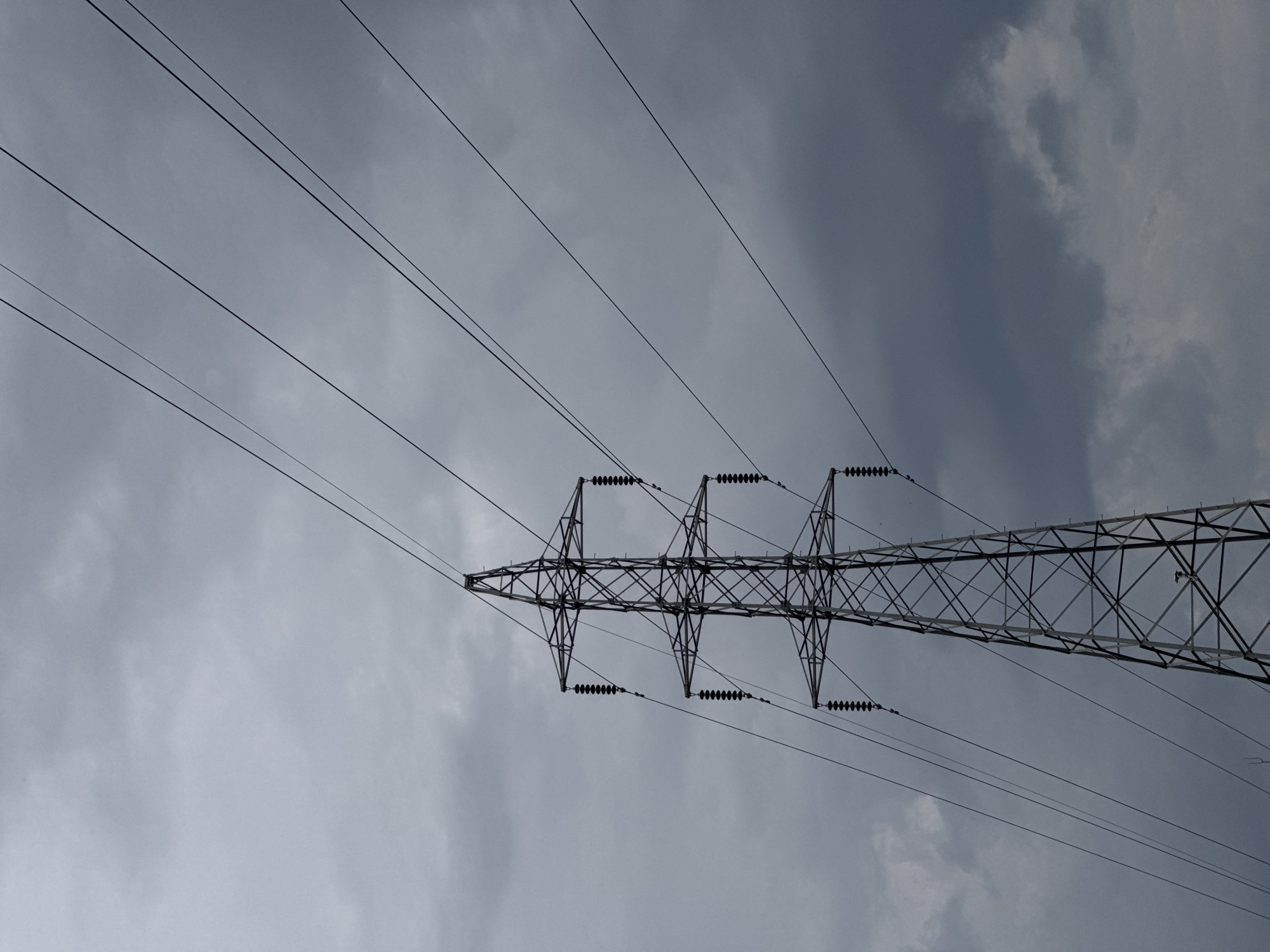 The image shows a transmission tower and power lines against a cloudy, overcast sky. The tower is constructed of metal and appears to be a standard lattice design. The power lines extend from the tower, running diagonally across the frame. The background is a uniform gray sky with varying cloud formations. The photograph is taken from a low angle, looking upwards at the tower. The provided location in บ้านกาญจน์กนก 23, Thailand is likely the location the image was taken, although no specific identifying features from that city are visible in the image.