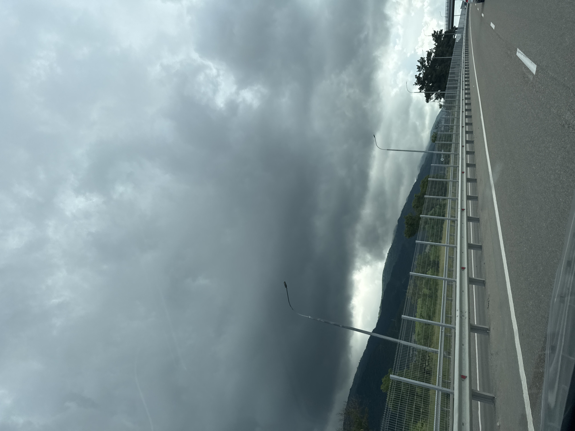 The image depicts a highway scene under a cloudy sky. The highway has multiple lanes, marked with white lines. A metal guardrail runs along the side of the road, separating it from a fenced area beyond. In the distance, mountains are visible, partially obscured by the dark, overcast clouds. There are streetlights positioned along the highway. The overall impression is one of a somber, possibly impending weather event. No people or specific vehicles are clearly discernible to identify the city of Kabardinka, Russia.