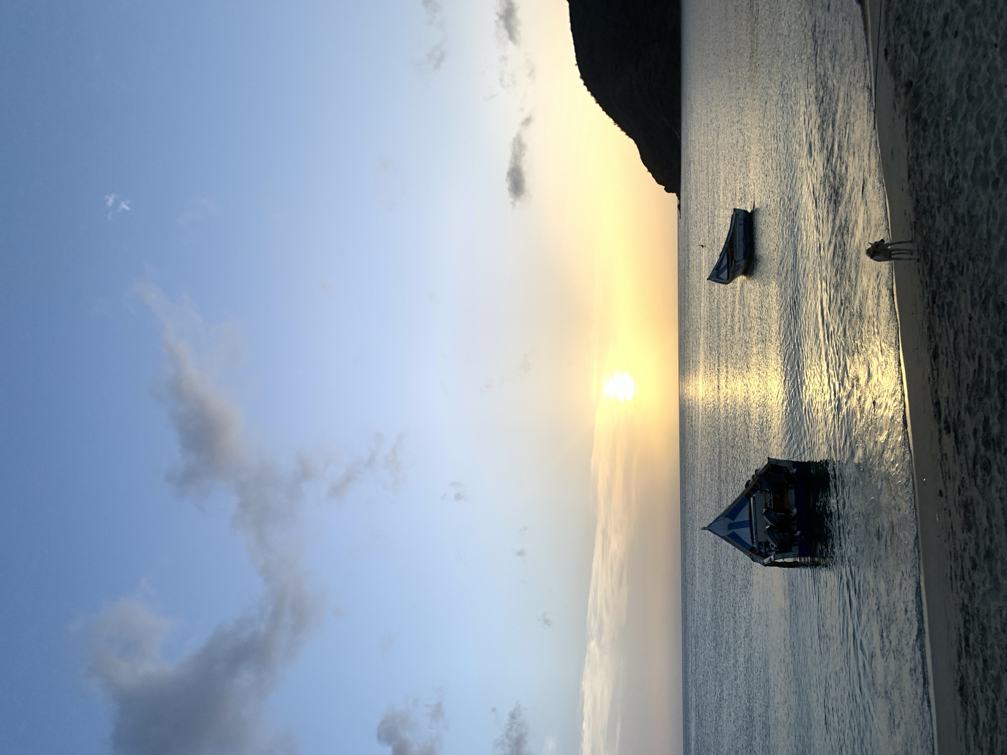 The image depicts a serene sunset scene on a beach in Juan Griego, Venezuela. The sky is a gradient of soft blues and yellows, with scattered clouds catching the last rays of the sun. Two small fishing boats are anchored in the calm, shallow waters of the sea. The boat on the left is closer to the shore and is painted with blue and white stripes. The boat on the right is further out and appears to be a lighter color. In the foreground, a sandy beach stretches along the water's edge. A lone dog, a light-colored mixed breed with a red collar, stands near the waterline, facing the sea. The silhouette of a dark, hilly coastline is visible in the distance on the right side of the frame. The overall atmosphere is tranquil and picturesque, characteristic of a peaceful evening in a coastal town.