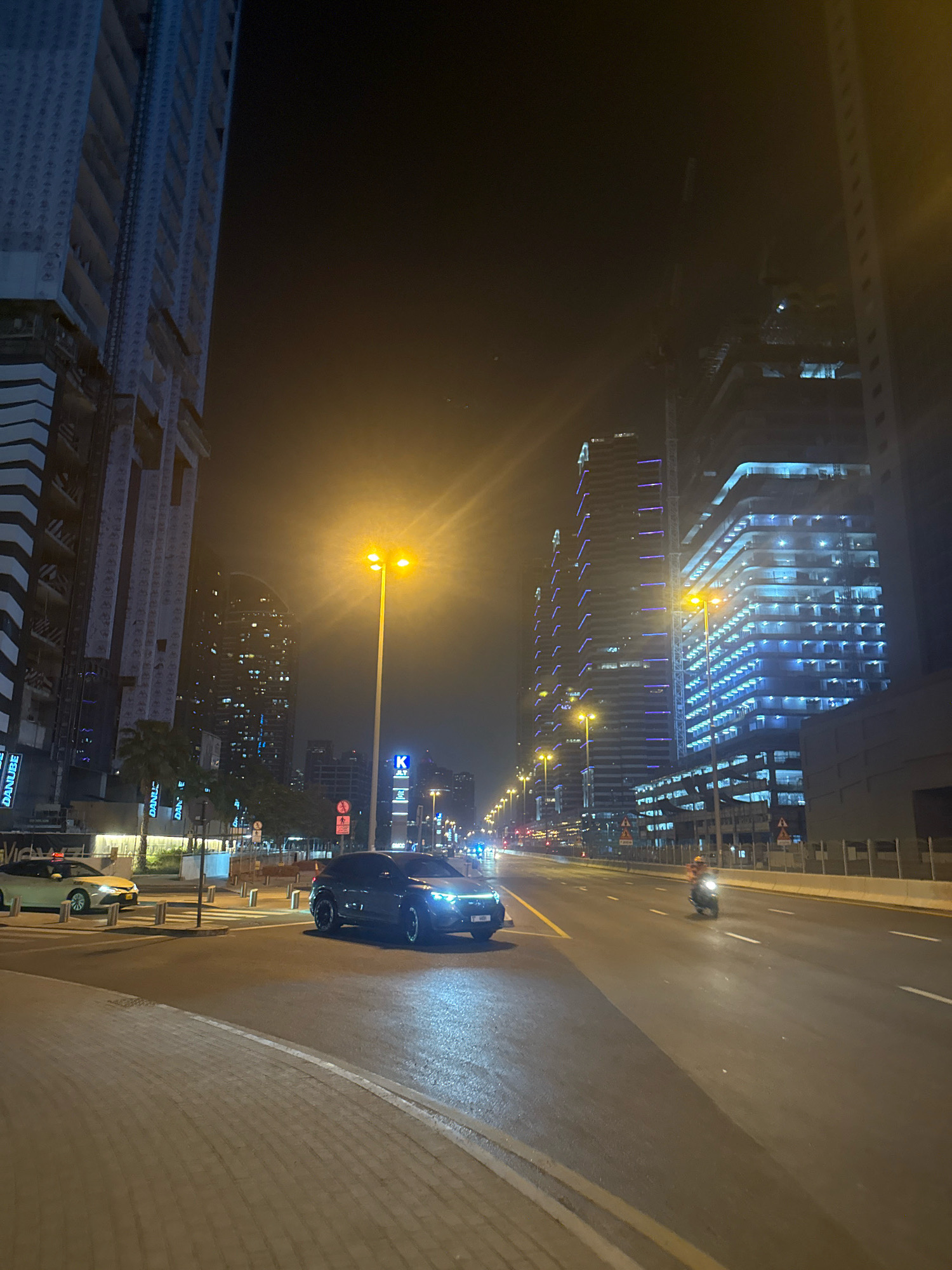 The image captures a nighttime street scene in Dubai, United Arab Emirates, likely on Sheikh Zayed Road, characterized by its towering skyscrapers and busy traffic. In the foreground, a dark-colored SUV is parked on the right side of the road, its headlights illuminated. Further down the street, a motorcycle with a rider is in motion, heading away from the camera. The left side of the road features a sidewalk made of textured concrete paving stones.
Dominating the background are several imposing skyscrapers, some of which are under construction, indicated by visible cranes and scaffolding. The buildings are adorned with vibrant blue and purple LED lighting, creating a striking visual contrast against the dark night sky. Streetlights with bright yellow bulbs line the road, casting pools of light and contributing to the overall illumination of the urban landscape. Traffic signals and various road signs are also visible along the thoroughfare, suggesting an active and regulated traffic flow. A white taxi is stopped on the left side of the road, near a pedestrian crossing. The overall impression is one of a modern, dynamic, and illuminated city at night.