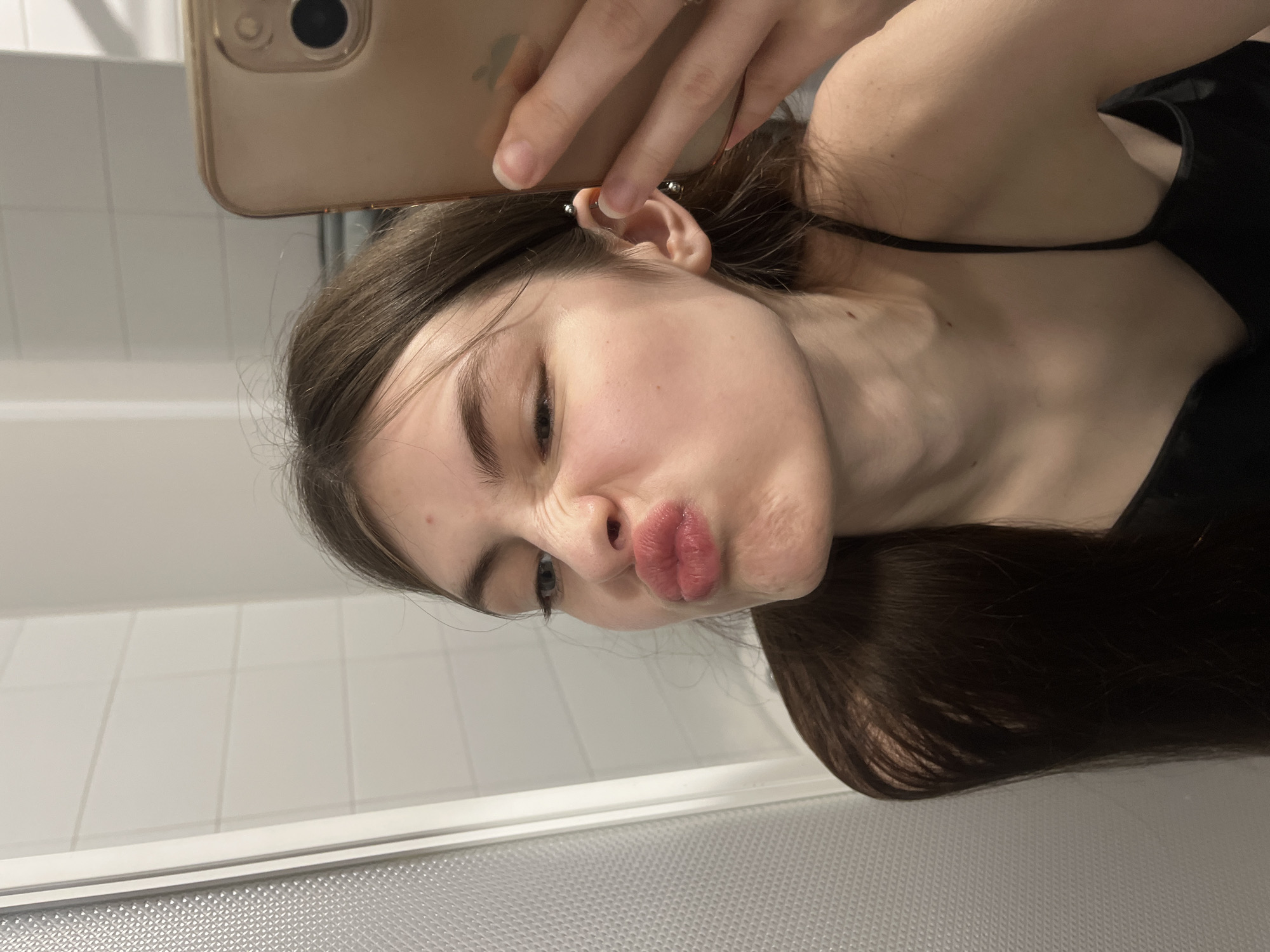 A young woman is taking a selfie in a bathroom with white tiled walls and a white ventilation grill on the lower part of the frame. She is holding a smartphone with a gold case in her right hand. The woman has brown hair and is wearing a black top with spaghetti straps. She is making a kissing face toward the camera. An industrial piercing is visible in her right ear. The location is not identifiable as Saint Petersburg, Russia from the image alone.