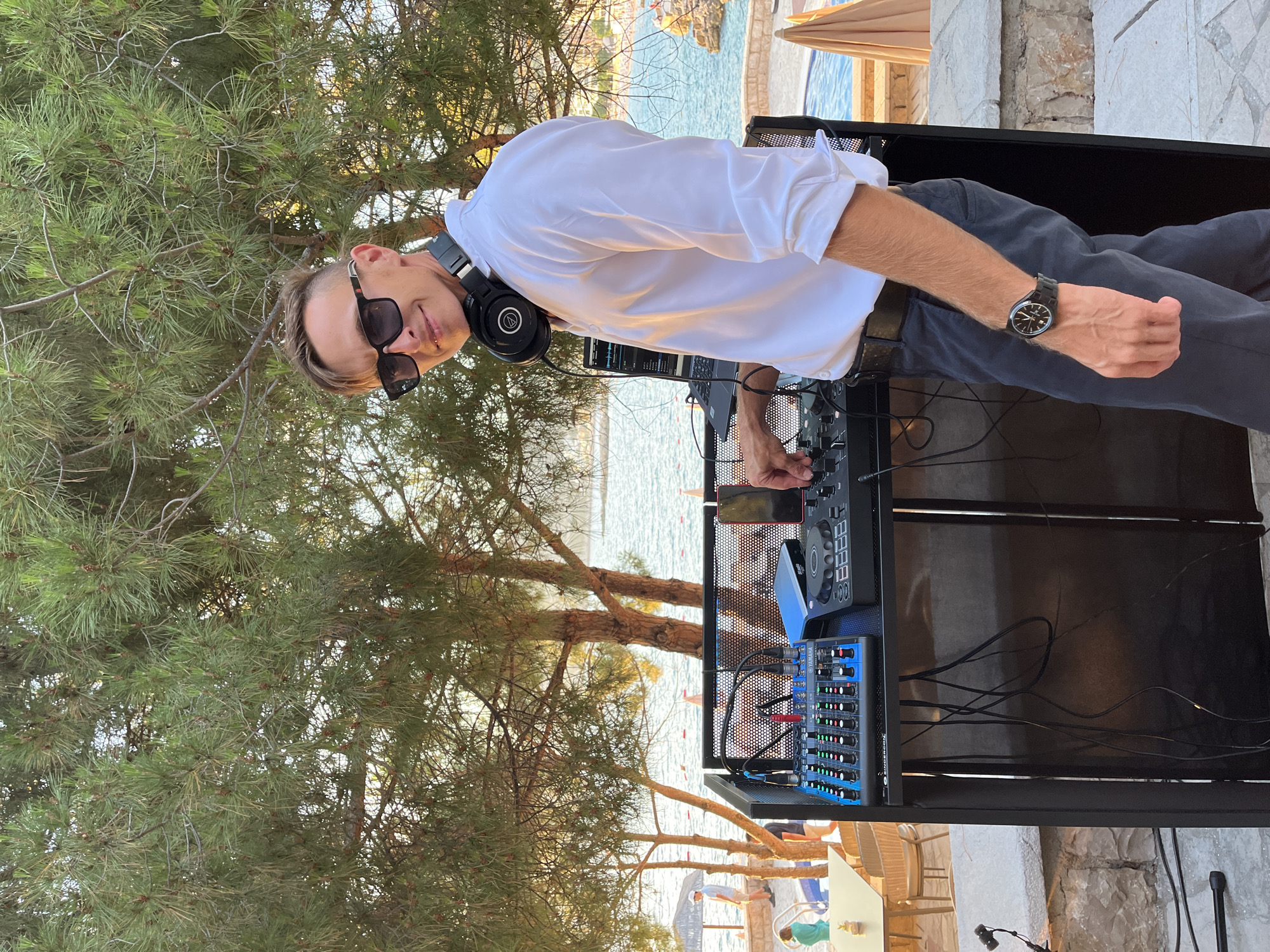 A man, identified as a DJ, stands behind a DJ setup on a terrace overlooking the sea in Pečurice, Montenegro. He is wearing sunglasses and a white button-down shirt, with headphones around his neck. The DJ setup includes a mixer, a laptop, and other equipment. In the background, there is a swimming pool and several trees. The overall scene suggests an outdoor event or party.