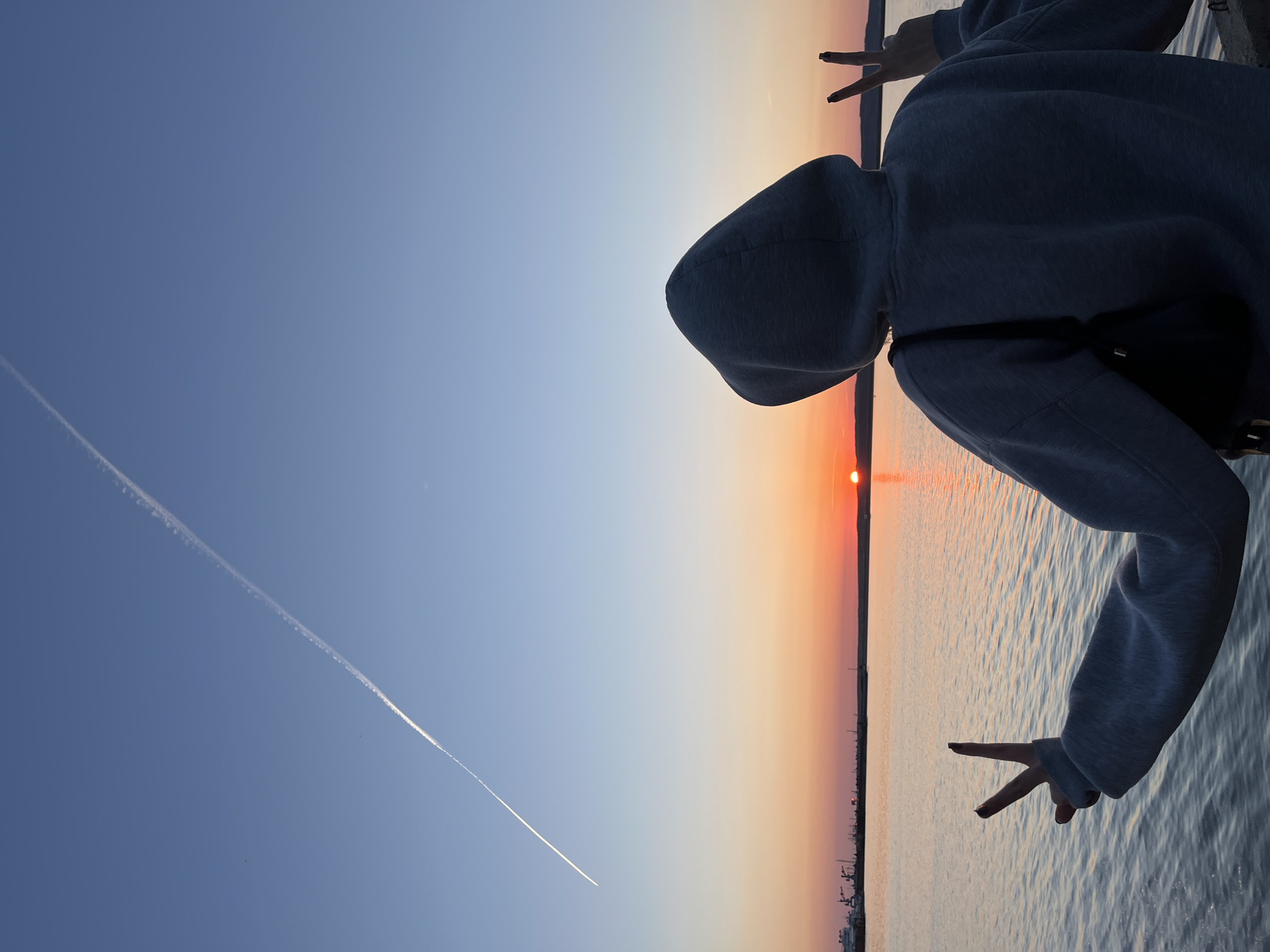 A person wearing a gray hoodie stands with their back to the viewer, with their hands outstretched making peace signs. The setting appears to be sunset over a body of water, likely a river or reservoir. The location is Chaikovsky, Russia, as indicated by the presence of industrial structures and cityscape along the shoreline. A contrail is visible in the sky to the left. The sun is setting on the right, with a gradient of orange and red tones across the sky and reflected in the water. The scene seems to depict an individual enjoying the sunset, framed by their silhouette and gestures.