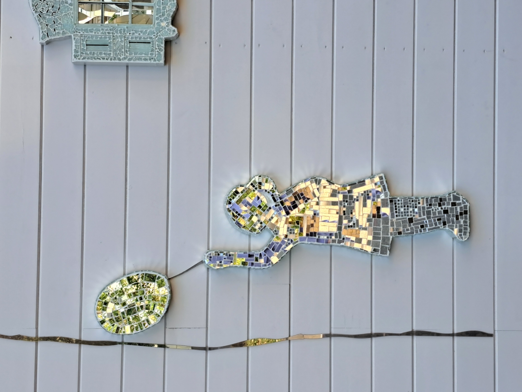 A mosaic depicts a young person holding a balloon, affixed to a light gray wooden wall. The mosaic is composed of numerous small, irregular mirror shards. The balloon, an oval shape, is positioned above and to the left of the figure. A thin, dark vertical line, also made of mosaic pieces, extends from near the balloon downwards, partially to the left of the figure, resembling a string. The figure itself is silhouetted, with the mosaic shards reflecting greenery, sky, and building textures, creating a fragmented image. To the upper right of the main mosaic, a separate, smaller mosaic piece in a light blue-green hue suggests a window frame with a divided pane, also made of reflective materials. The wall is constructed of horizontal planks. The image does not provide information about a specific location in Irkutsk, Russia.