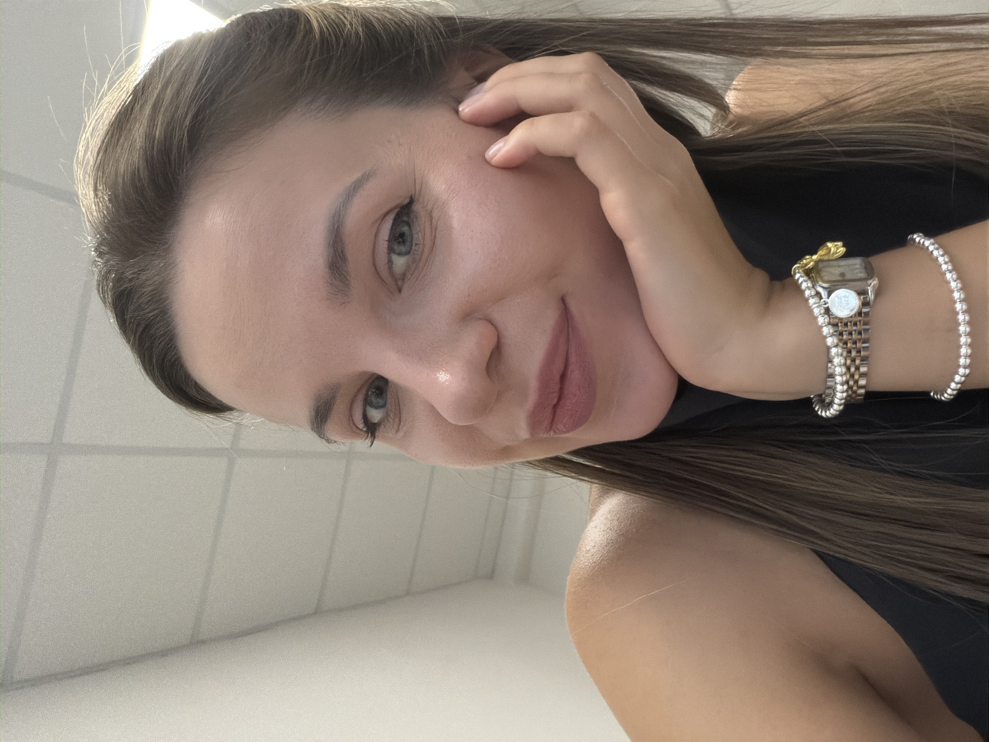 The image shows a close-up, selfie-style shot of a woman resting her chin on her hand. She has light skin, blue eyes, and brown hair pulled back. She is wearing a black top and a bracelet with charms. The background appears to be an indoor environment with a white wall and a tiled ceiling, possibly an office or public building. There is no indication of the city of Homyel, Belarus.
