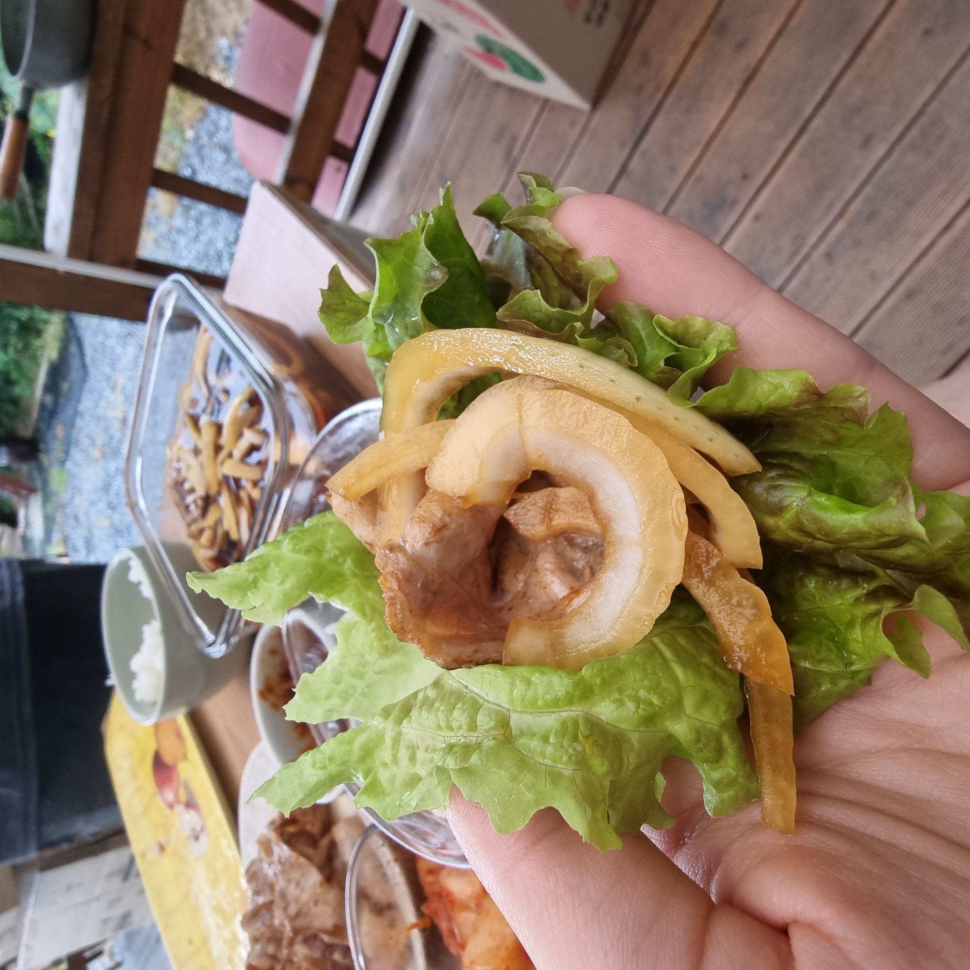A person is holding a lettuce wrap filled with pork and pickled radish, part of a meal in Dunnae-myeon, South Korea. The wrap is held in the palm of a hand, showcasing the vibrant green of the lettuce, the tender-looking pork, and the translucent, yellowish slices of pickled radish. In the background, a wooden deck suggests an outdoor setting, possibly a patio or a porch. Various dishes are visible on a table, including a glass container filled with what appears to be seasoned fried noodles or similar side dish, small bowls with sauces or kimchi, and a cup of white rice. The lighting suggests it is daytime. The overall scene evokes a casual meal, likely outdoors, enjoying traditional Korean cuisine.