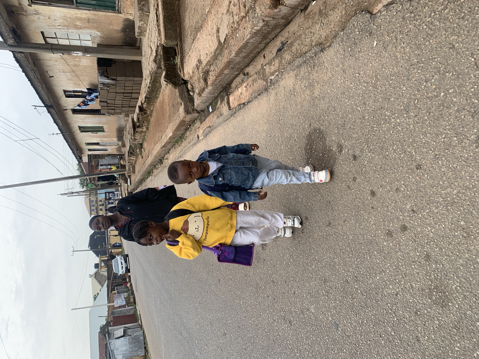 The scene shows a family of three walking down a paved road in what appears to be the city of Jos, Nigeria. An adult female, dressed in a black outfit with red accents, walks alongside a young girl in a yellow sweater with a cartoon print. The girl carries a small purple bag. A young boy wearing a denim jacket and light-colored pants walks slightly ahead. The background features weathered buildings and infrastructure, including overhead power lines and a few visible shops. The setting suggests an urban or semi-urban residential area.
