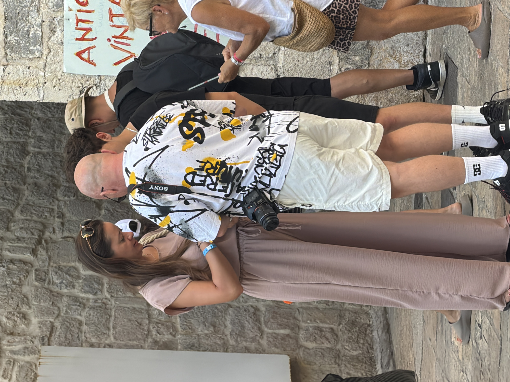 In Budva, Montenegro, a group of four individuals is captured in a street scene. A woman in a mauve jumpsuit and sunglasses on her head is engaged in conversation with a bald man. The man is wearing a white t-shirt with a black and yellow abstract print and holding a camera around his neck. To his right, another man in a black t-shirt and a khaki hat faces away from the camera. On the far right, a woman with blonde hair and glasses is looking at the man with the camera and carrying a woven bag. Behind the main group, a sign with red lettering reads "ANTIC VINTAGE". The background consists of a stone wall, and the ground is paved with cobblestones.