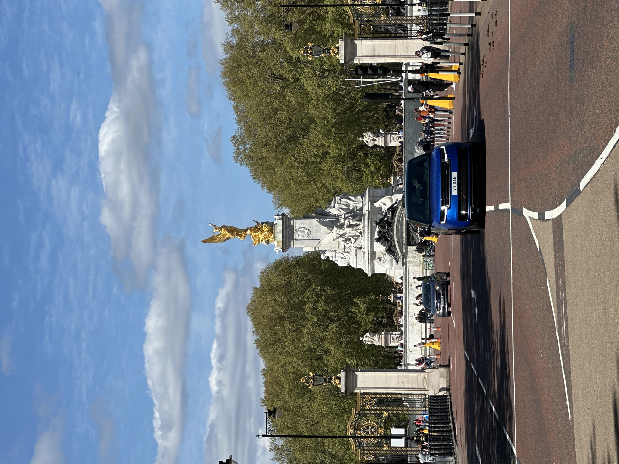 The image depicts a scene on The Mall in London, United Kingdom, with Buckingham Palace in the background. A blue vehicle with the license plate "V7 DAN" is driving on the road. Pedestrians are visible behind a barrier along the palace fence. A large, gold statue tops a monument, surrounded by white statues. The sky is blue with white clouds, and tall trees line the road. Traffic lights and street lamps are also visible.