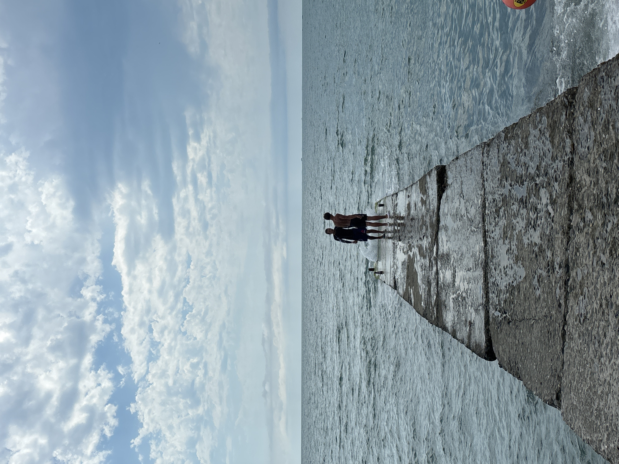Two young men stand on a concrete pier jutting into a calm, grey sea under a cloudy sky. The pier is wet and appears to be old, with visible cracks and seams in the concrete. The men are facing away from the camera, looking out at the water. One man is wearing a dark long-sleeved shirt and shorts, while the other is shirtless and wearing shorts. They are positioned near the end of the pier. In the distance, the horizon line is visible, separating the sea from the sky. The sky is filled with a mix of white and grey clouds. The scene is serene and appears to be a quiet moment by the sea. The location is the city of Sochi, Russia, likely a public pier or breakwater accessible to swimmers or beachgoers.