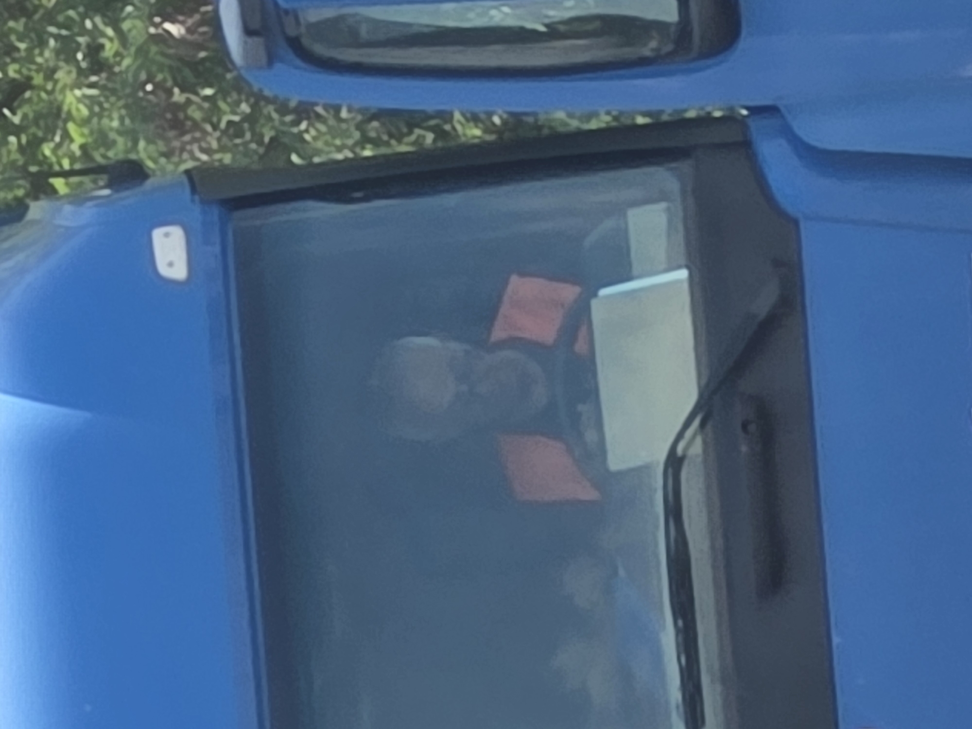 A man wearing a high-visibility orange vest sits in the driver's seat of a blue truck. He is looking at a laptop that is on the dashboard. The truck is parked next to another blue truck. Trees and foliage are visible in the background. The image appears to have been taken in Frankfurt, Germany.