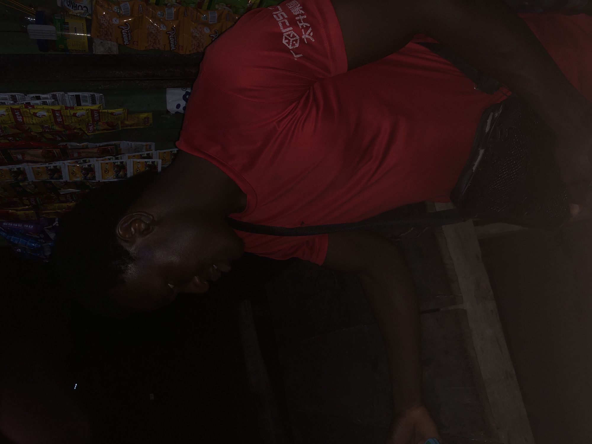 The image depicts a young Black male wearing a red t-shirt and a black satchel, standing in what appears to be a small shop or market stall in Ado, Nigeria. The background is filled with various packaged goods, likely food items, hanging from the walls. The lighting is dim, suggesting it might be evening or an indoor setting with limited illumination. The man is positioned to the right of the frame, looking downwards and to his left. His facial features are partially obscured by the shadow and his downward gaze. The overall scene conveys a candid moment, possibly at a local convenience store or a similar retail environment.