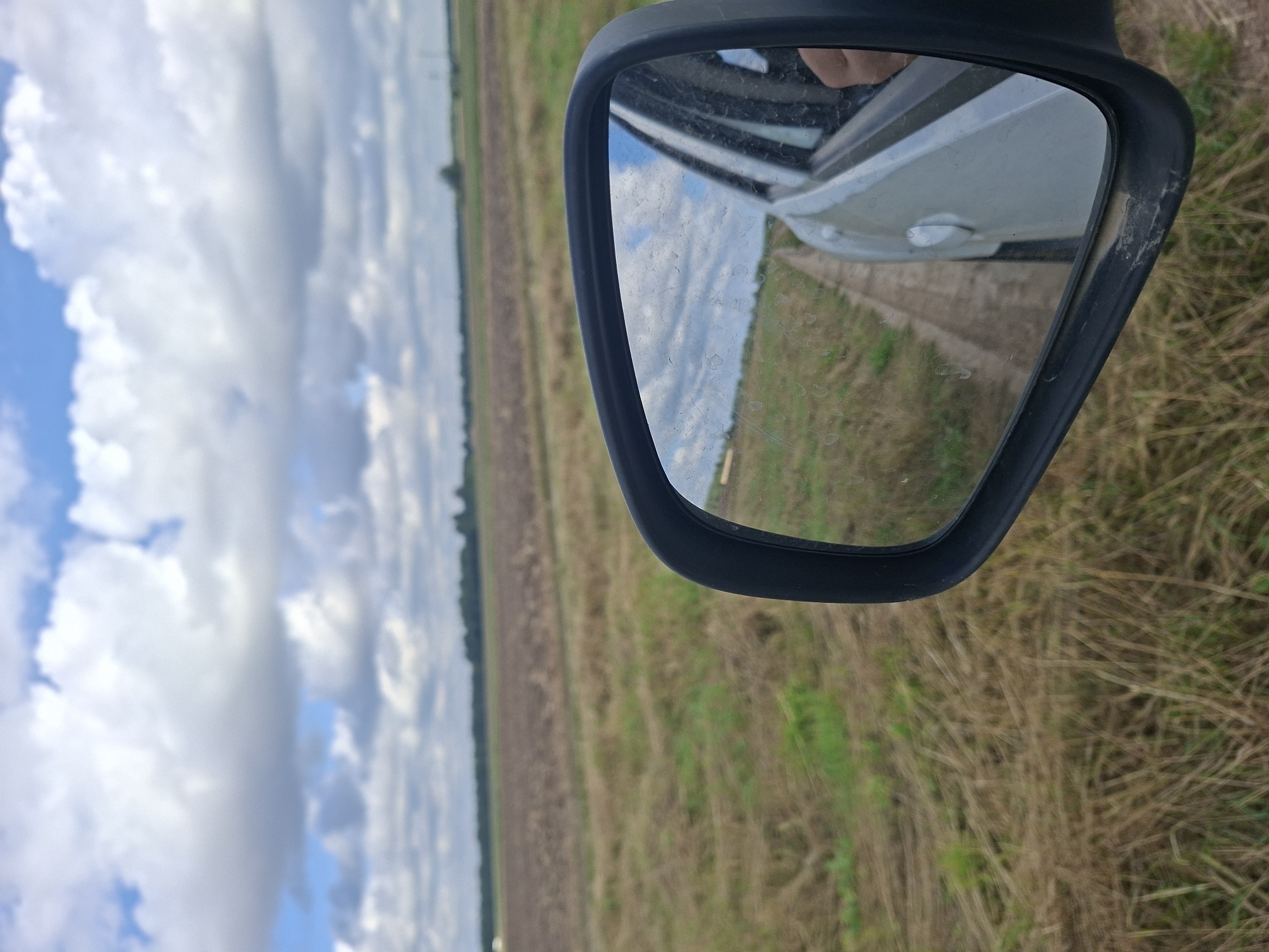 A side-view mirror of a white car reflects a cloudy sky and a dirt road. The car is traveling through a rural area with a dry grassy field to the left and a harvested or tilled field in the distance, with a line of trees on the horizon. The area appears to be the Славенскі сельскі Савет, Belarus.